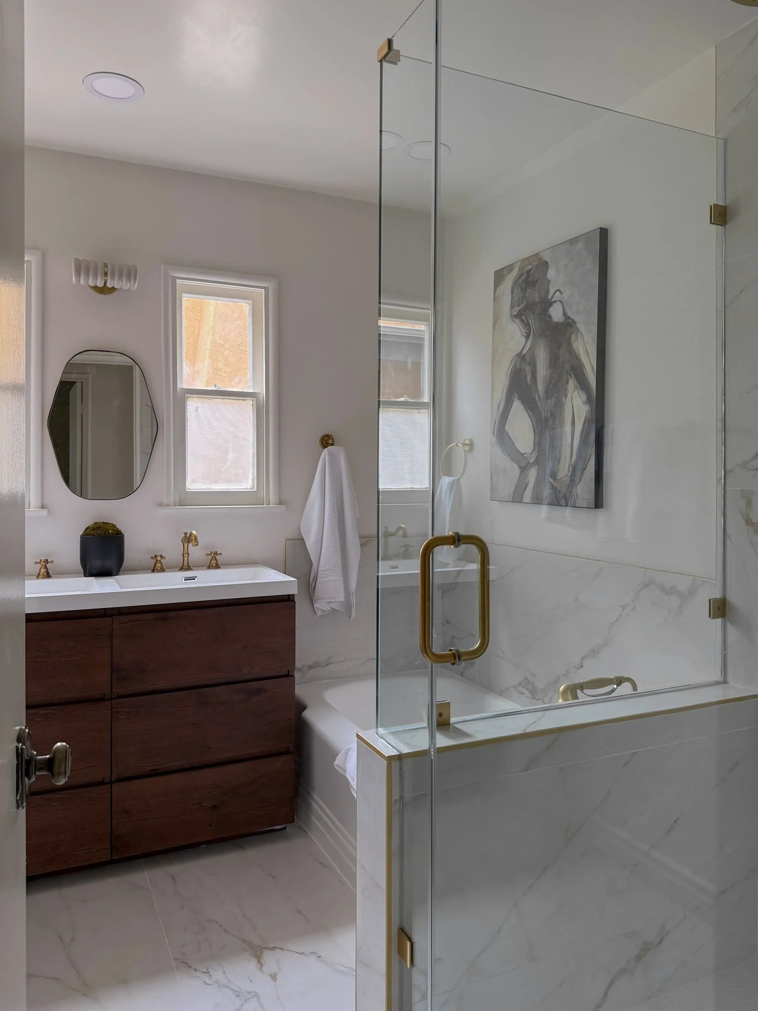 A modern bathroom with a wooden vanity, white sink, gold fixtures, a marble bathtub and shower enclosure, two windows, and contemporary artwork on the wall.
