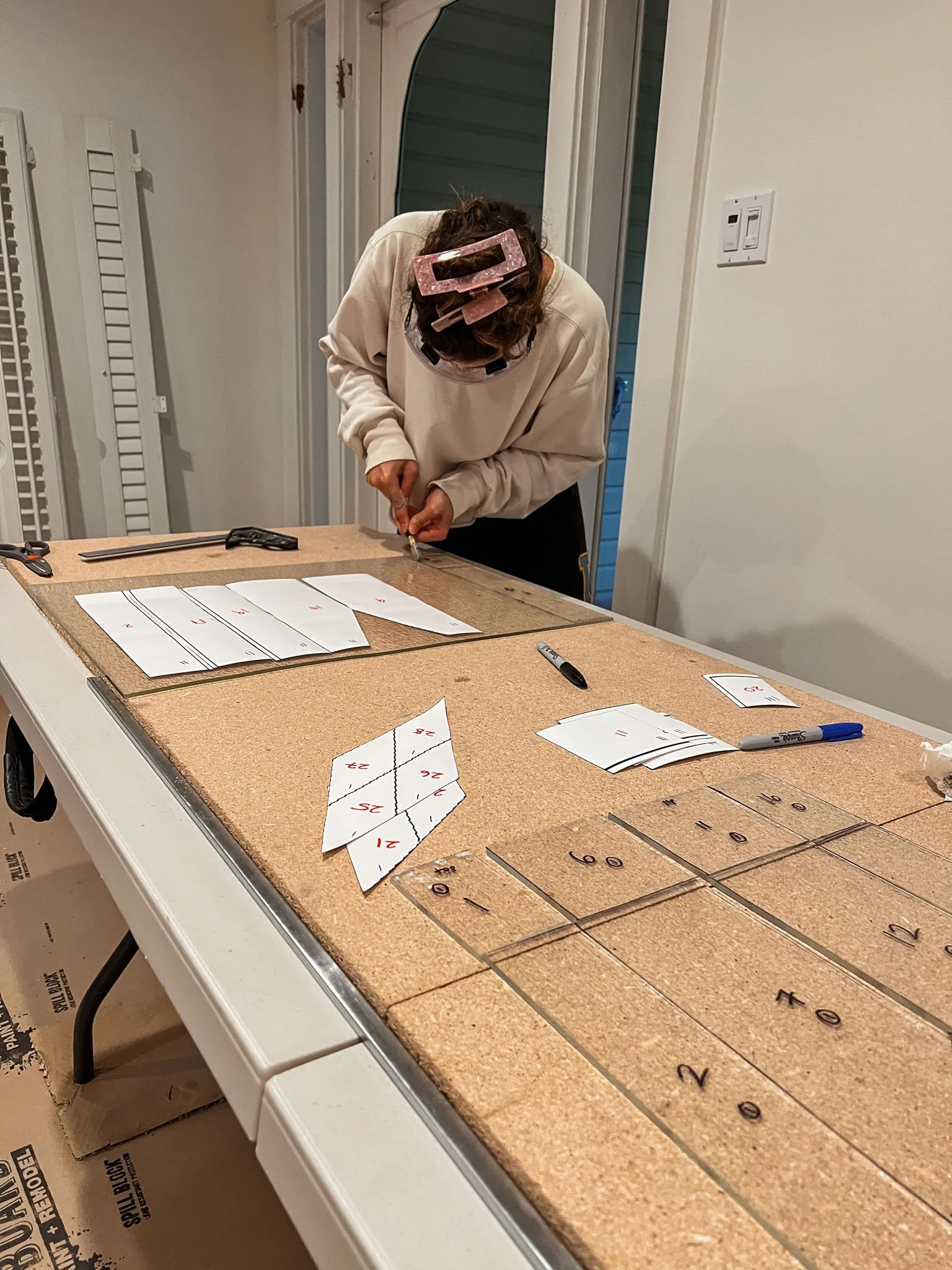 Person working on a woodworking project at a table with numbered and divided paper templates, wearing protective glasses, and using a small tool.