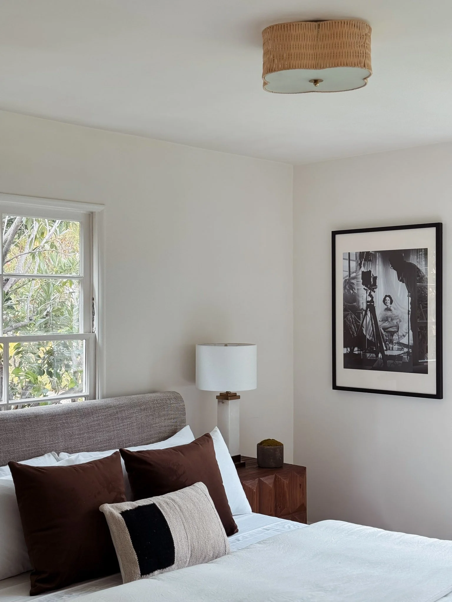 A bedroom with a bed that has white sheets and brown pillows, a wooden bedside table with a lamp, a black-and-white framed photograph on the wall, a window showing plant foliage outside, and a ceiling light fixture.