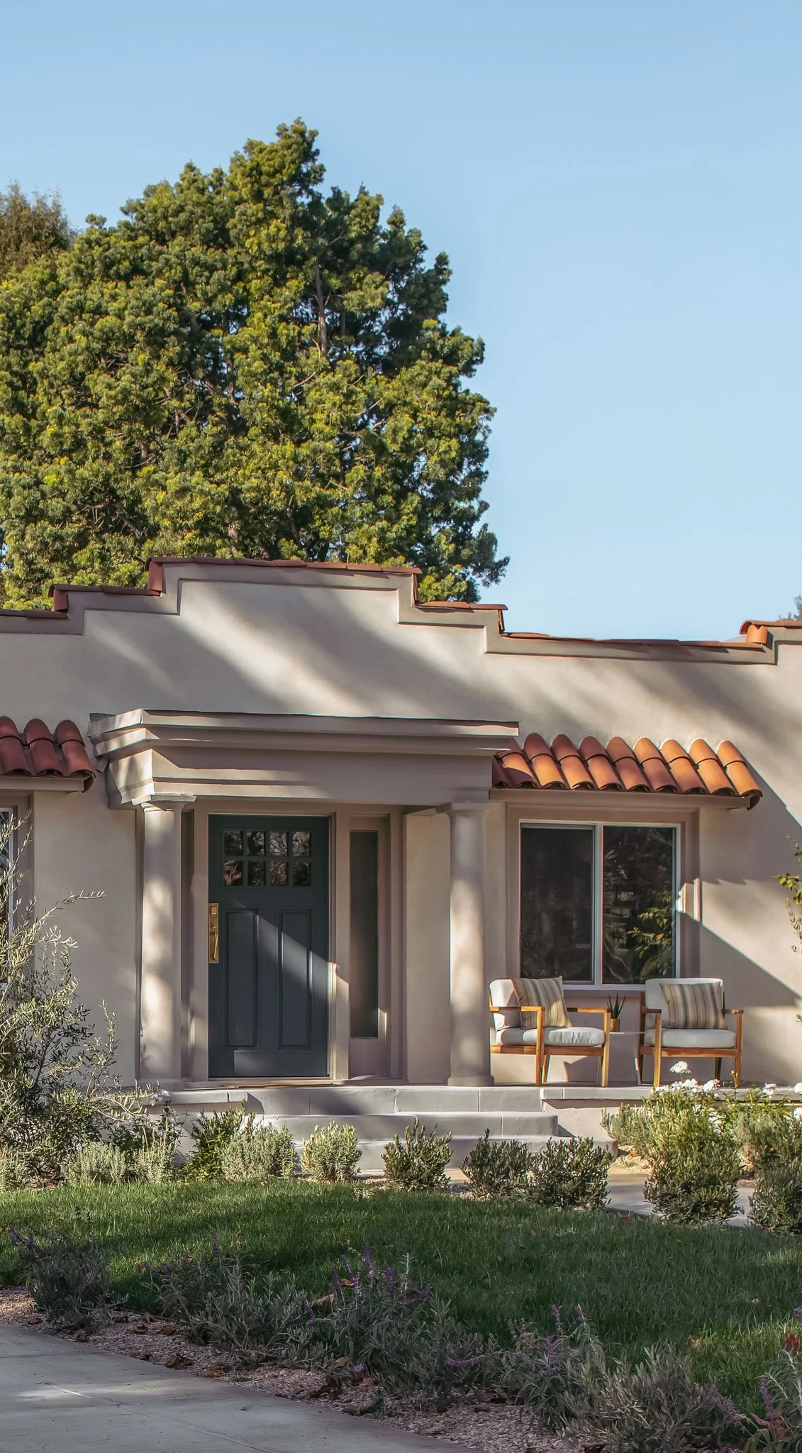 Front view of a house with a dark green door, two chairs on the porch, and a large tree in the background.
