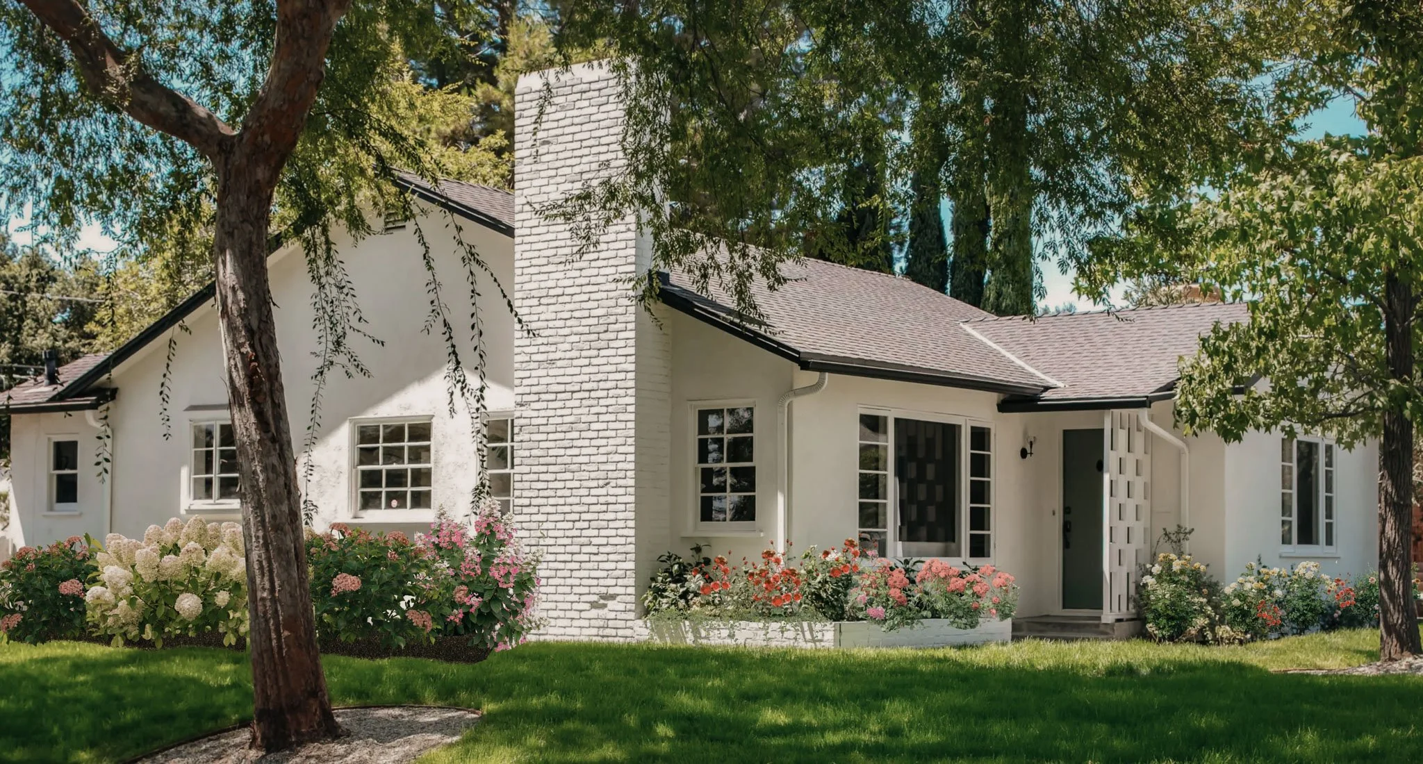 White house with a brick chimney, surrounded by colorful flowers and green trees, on a sunny day.