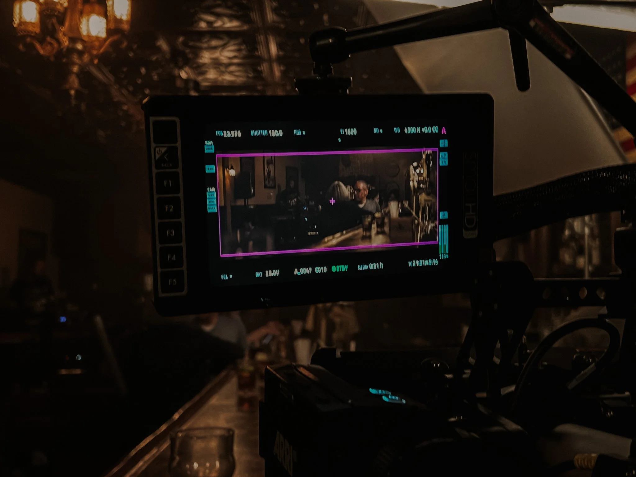 On a film or camera monitor, a dimly lit scene of a person in glasses sitting at a table in a restaurant or bar. Reflections and ambient lighting create a cozy atmosphere.