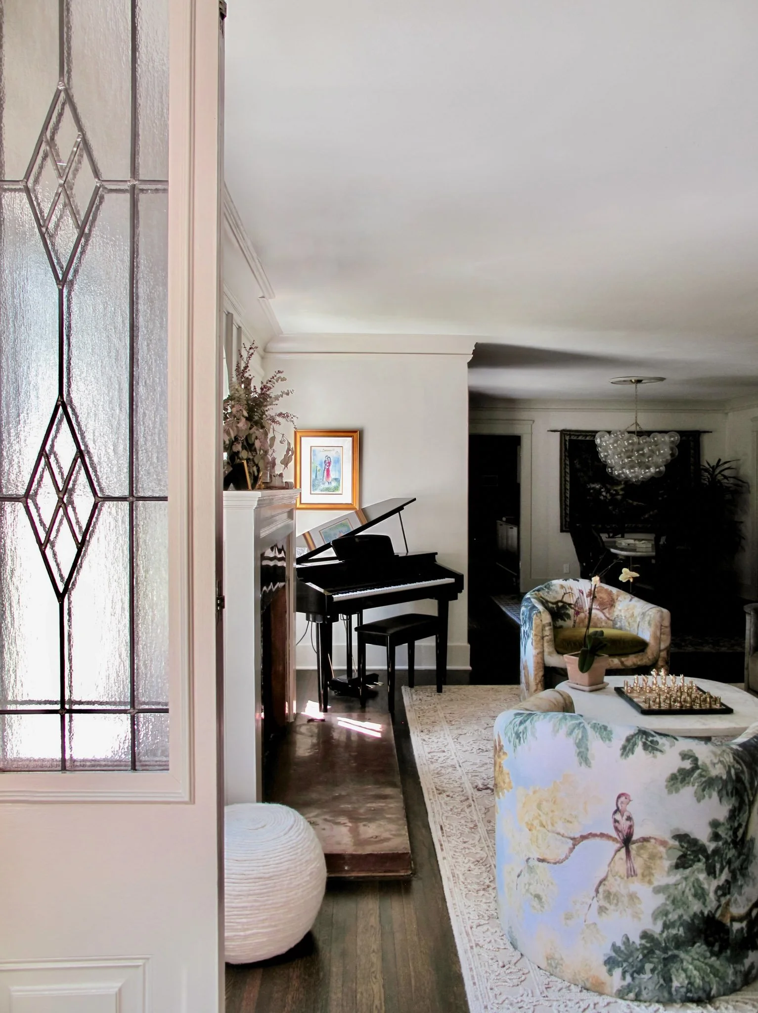 Living room with a black grand piano, floral upholstered armchair, sofa, and a glass chandelier.