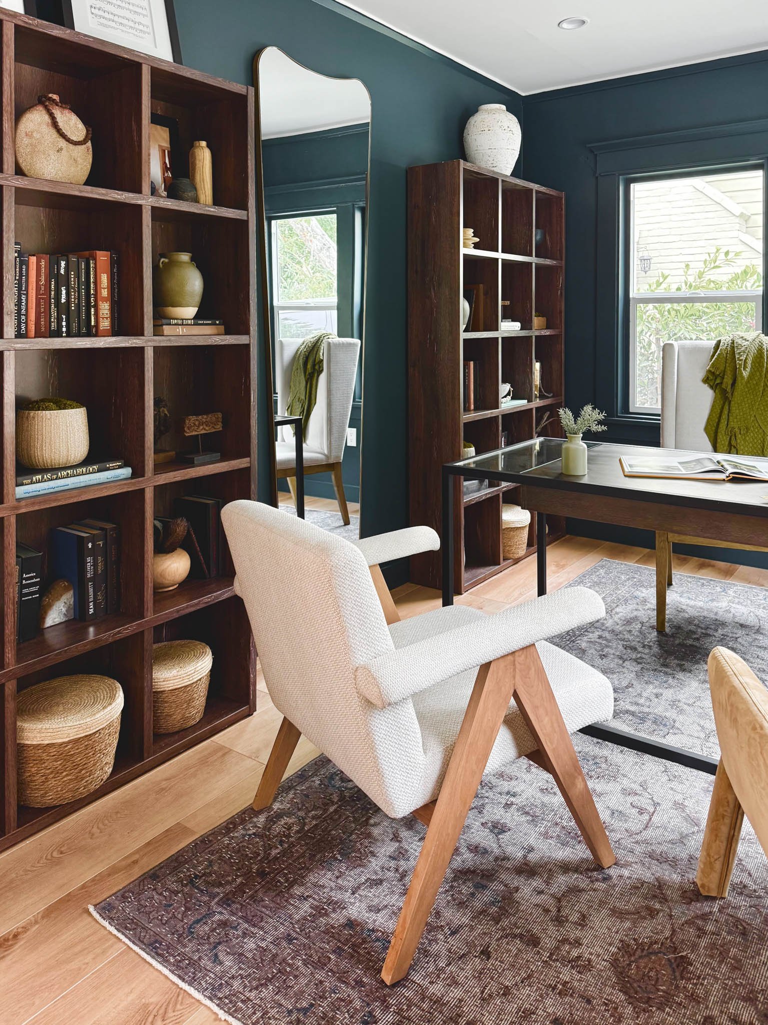 A cozy room with a white upholstered armchair with wooden angled legs, a dark wood table with an open book and a small vase with green plants, two dark wood bookshelves filled with books and decorative vases, a large mirror, and a window with plants 