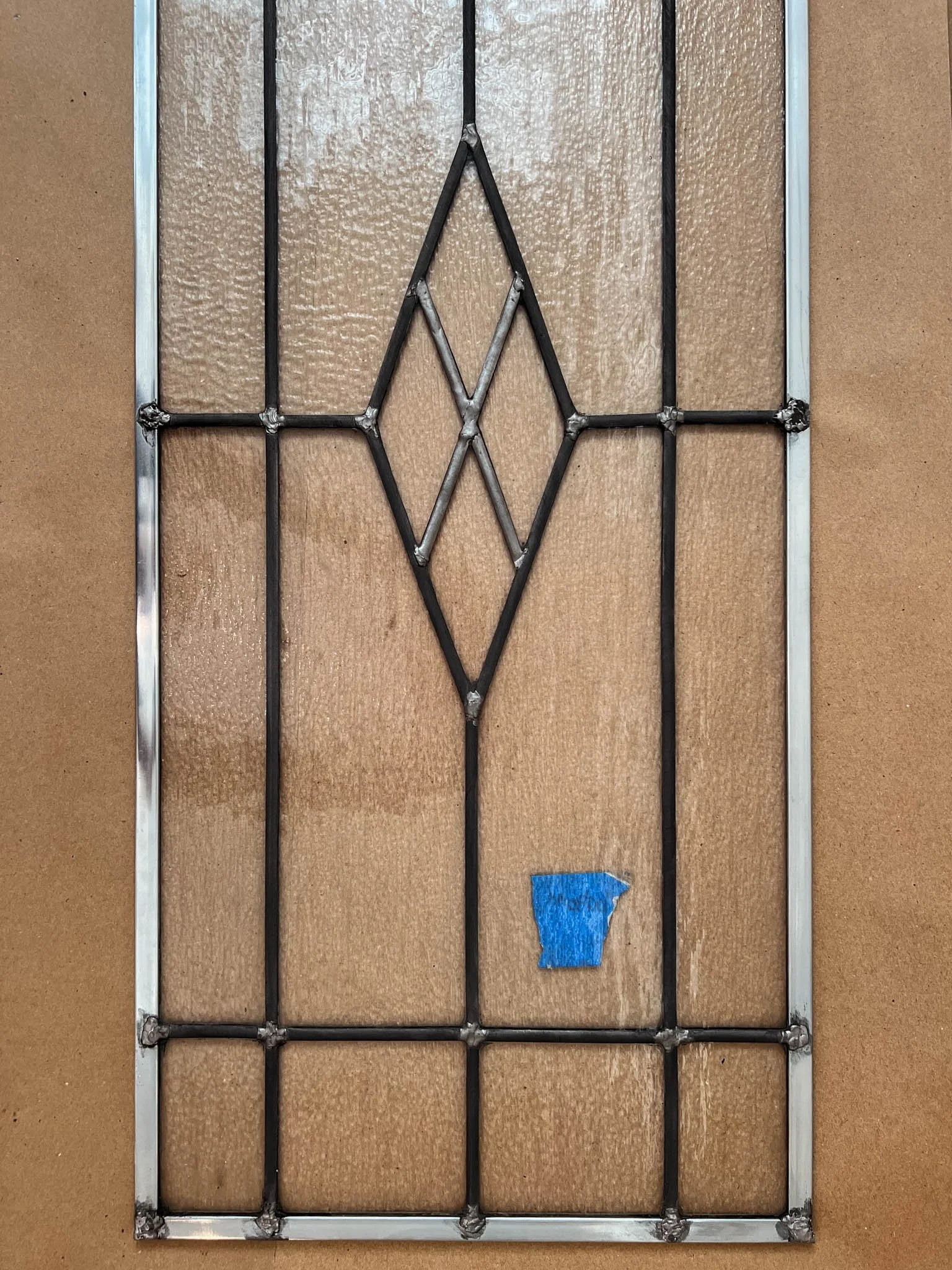 Metal window security grille mounted on a brown wall with a small piece of blue painter's tape on it.