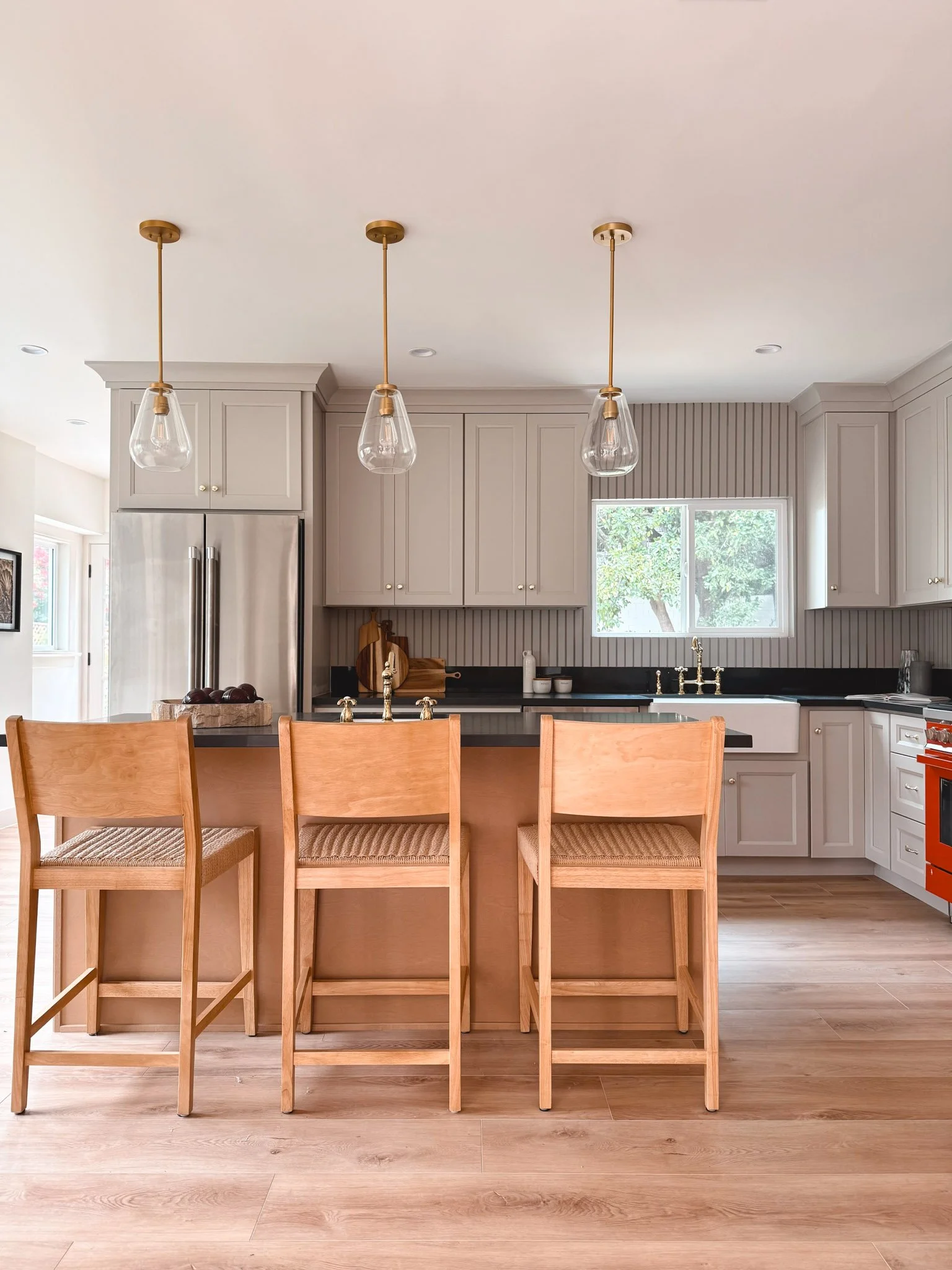 Modern kitchen with white cabinetry, black countertops, three pendant lights, wooden bar stools, stainless steel refrigerator, window with trees outside, and a farmhouse sink.