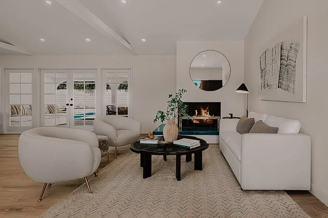 Modern living room with white sofa, two white armchairs, a black coffee table with books and a vase, a fireplace with a mirror above, a floor lamp, and a large artwork on the wall. French doors lead outside to a pool area.