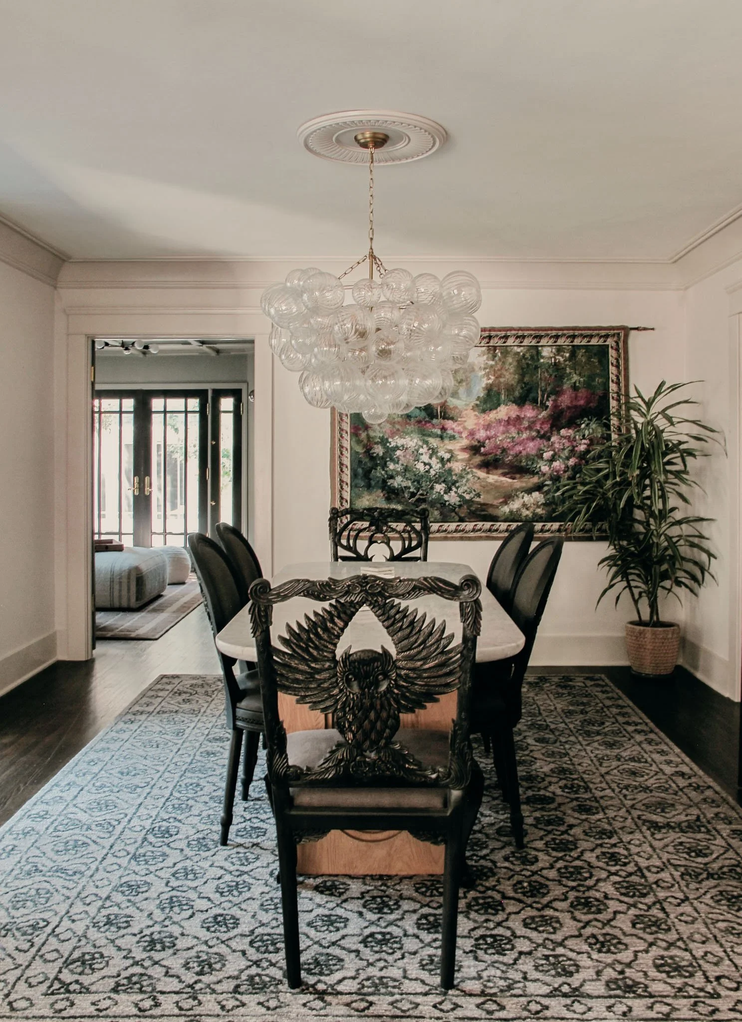 Dining room with a rectangular table surrounded by six black chairs, a chandelier hanging from the ceiling, a framed floral painting on the wall, a large potted plant in the corner, and a patterned rug on the dark wood floor.