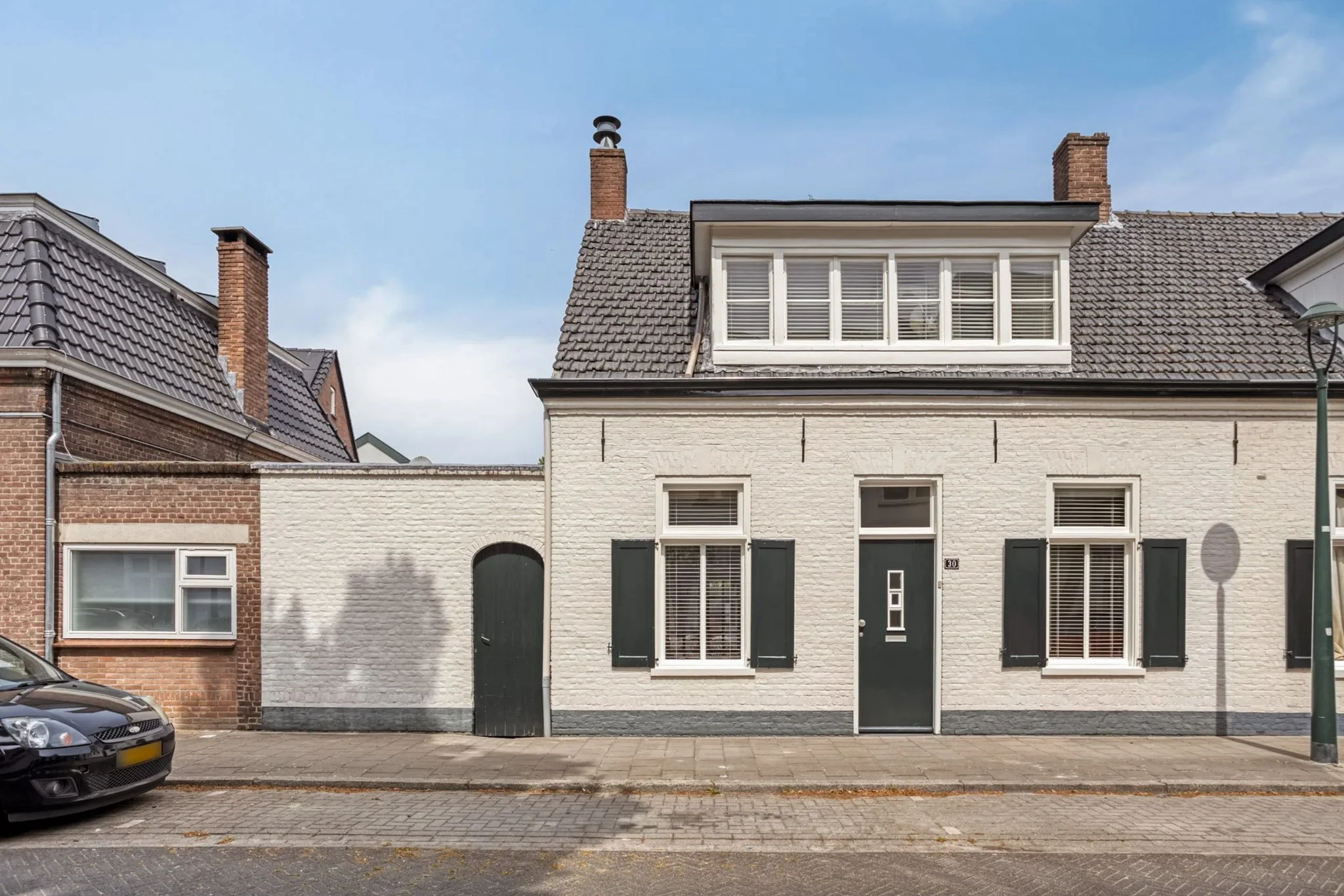 Street view of a white brick house with black shutters, a dark door with a glass panel, and a large dormer window on the roof. A shadow of a tree is cast on the wall, and a car is parked on the street.