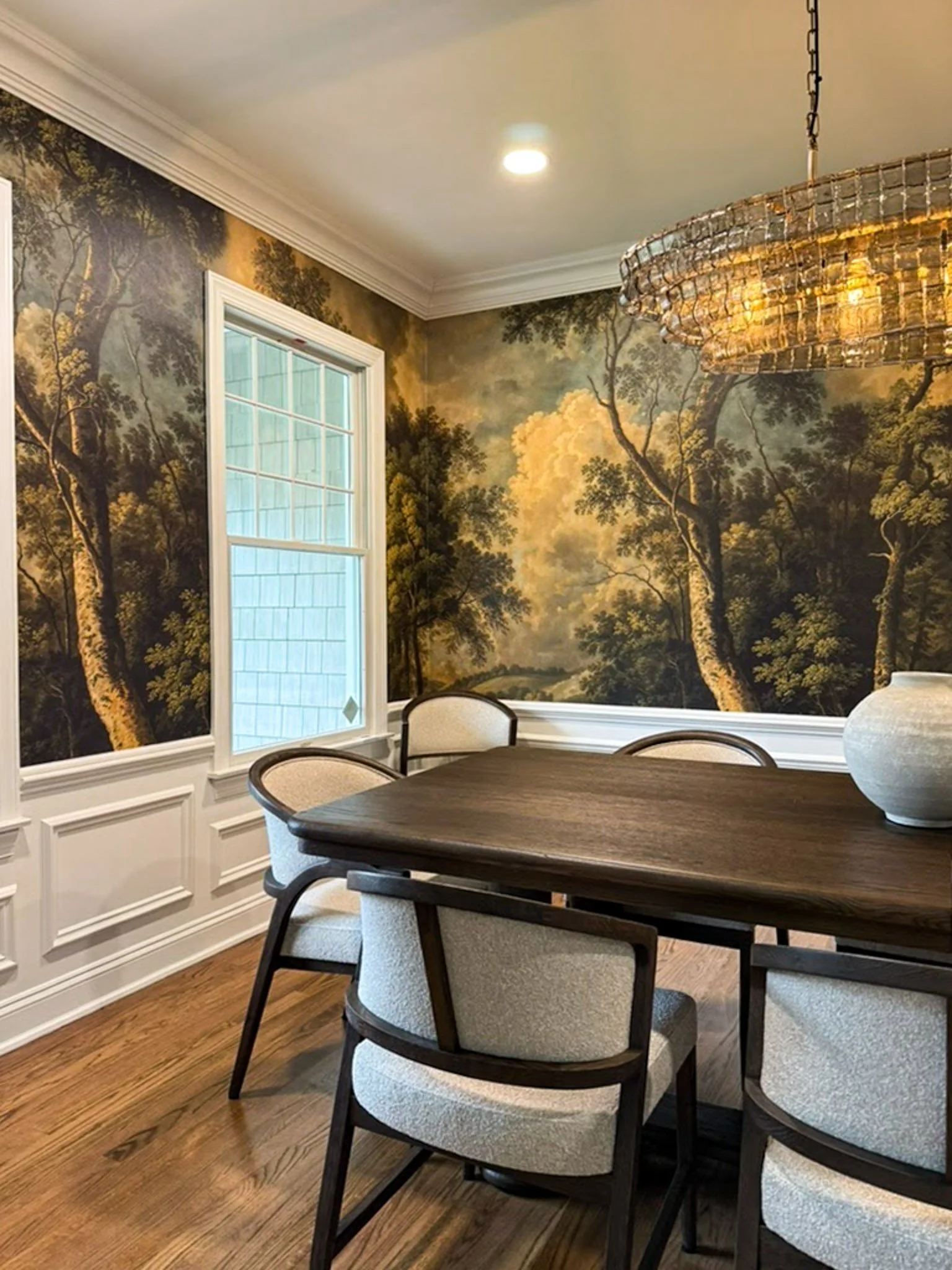 A dining room with a wooden table and four upholstered chairs, a large mural of a forest landscape on the wall, and a round chandelier hanging from the ceiling.