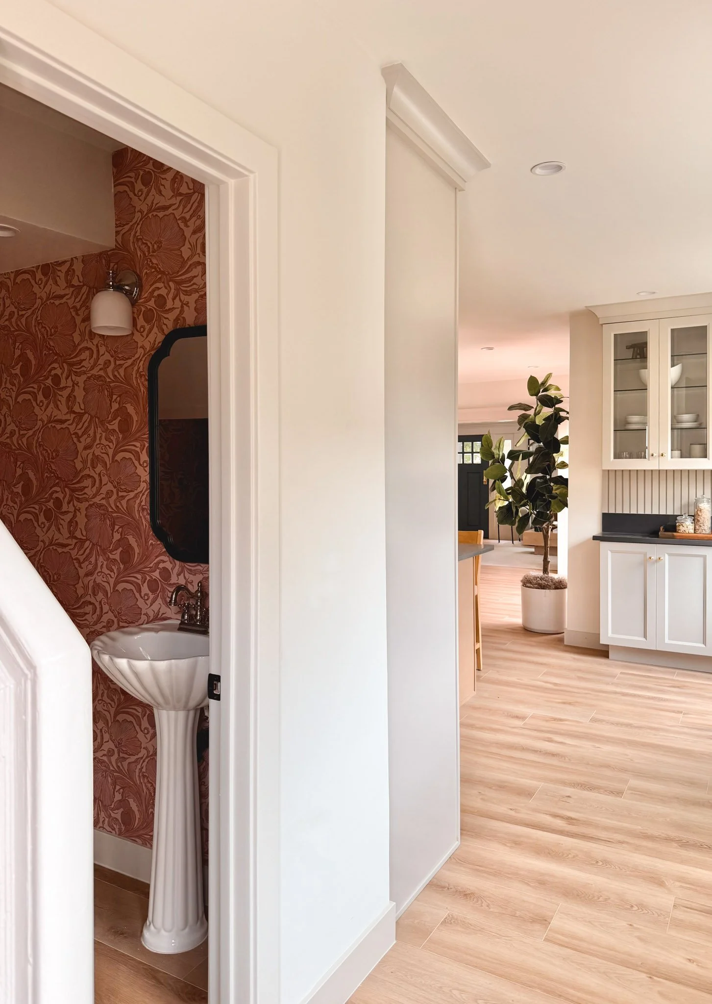 A partial view of a hallway in a home, showing a small bathroom with red floral wallpaper, a white pedestal sink with a black mirror above, and part of a kitchen with white cabinetry, a potted plant, and light wood flooring.