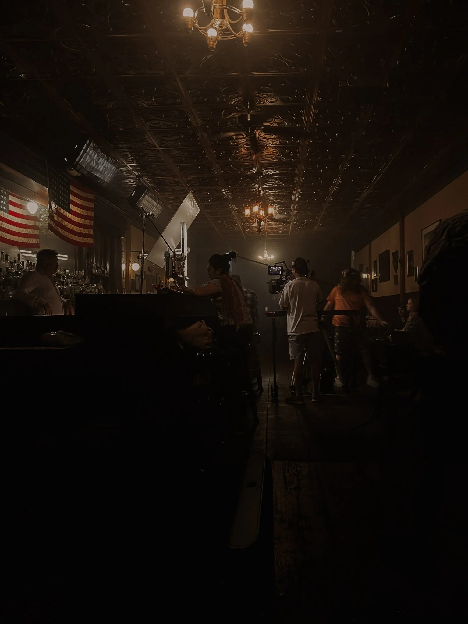 People filming a scene or recording in a dimly lit bar or restaurant with American flags hanging on the wall and a decorative ceiling.