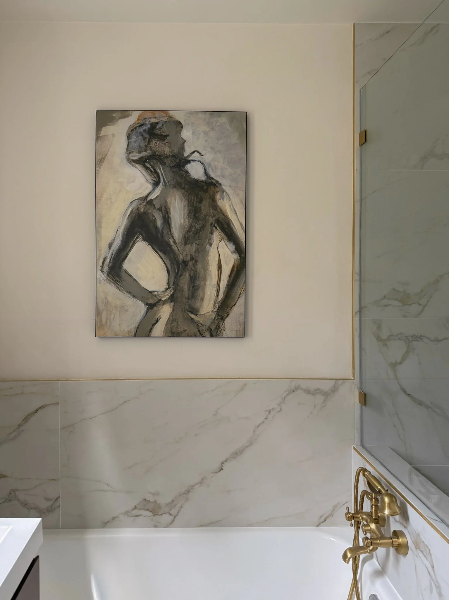 A modern bathroom with marble walls, a white bathtub, and gold fixtures. A painting of a nude woman with short hair stands on the wall.