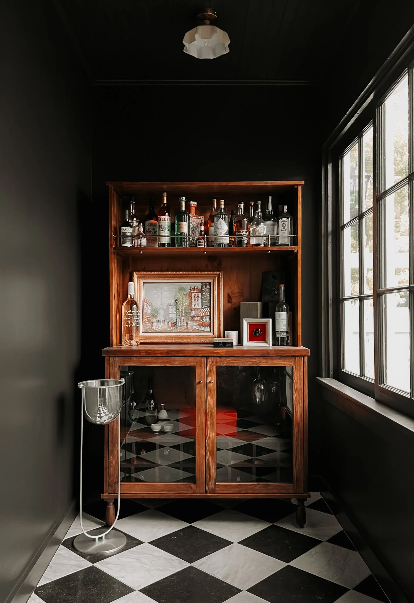 A vintage wooden bar cabinet with glass doors, filled with liquor bottles on the top shelf and some displayed artwork and decorative items on the lower section, next to a large window with black framing and a checkered black-and-white floor.