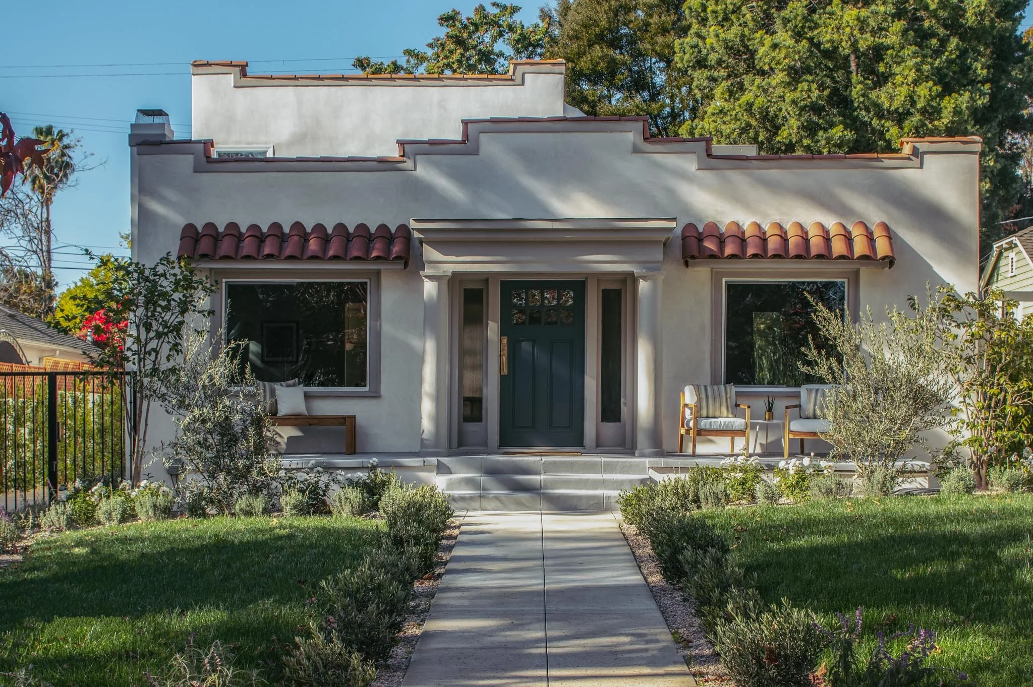 Front view of a modern house with white stucco exterior, red clay tiles on the roof, and a dark green front door. The house has a small porch with two chairs on the right side and a bench on the left, surrounded by a well-maintained garden with bushe