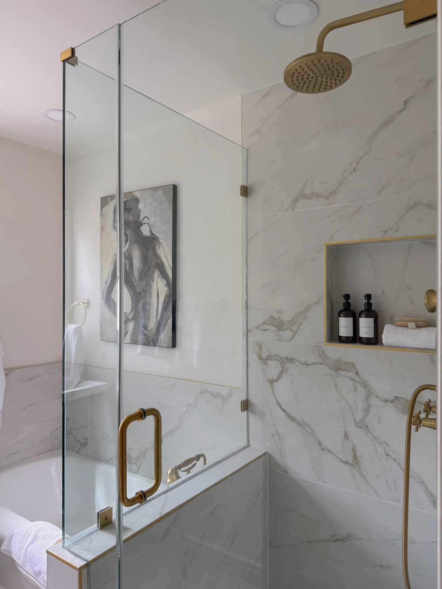 A modern bathroom shower area with marble walls, a clear glass door with a gold handle, a gold showerhead, and built-in shelves holding black bottles, a towel, and a hairbrush.