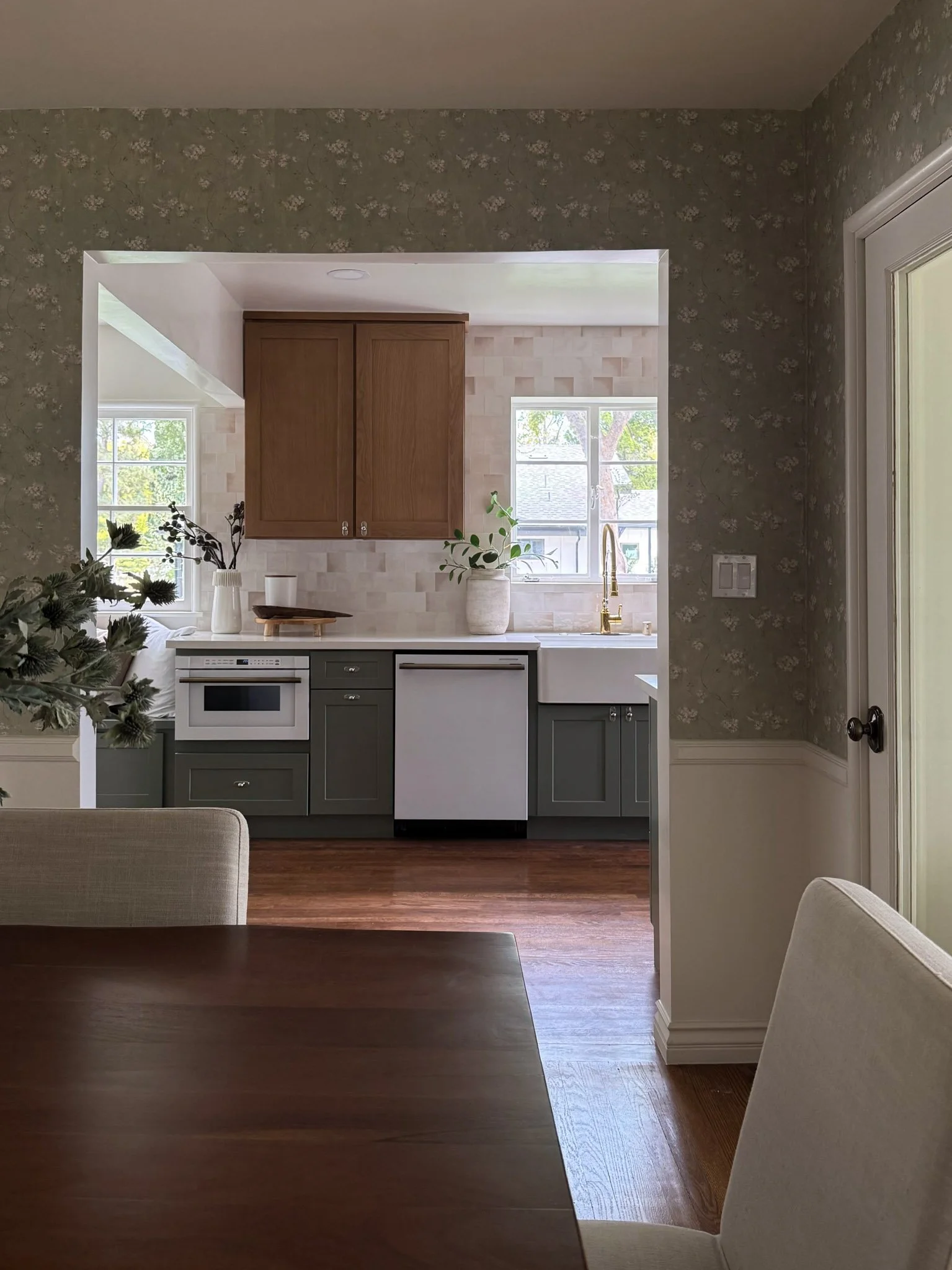 View of a kitchen with gray cabinetry, a white farmhouse sink, a window above the sink, and wood flooring, with a glimpse of a dining area with a wooden table and light-colored chairs.