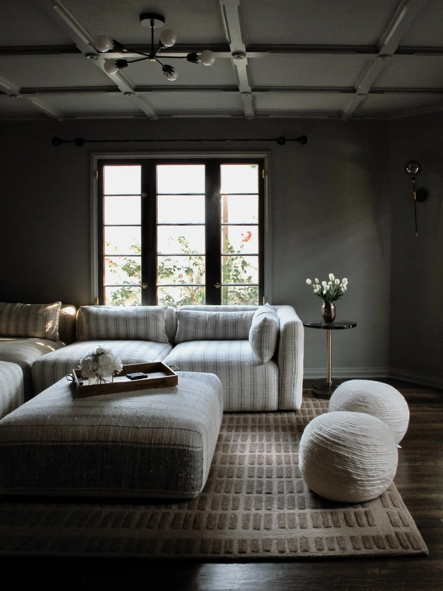 Living room with a large window, white patterned sectional sofa, ottoman, two textured round poufs, a side table with a vase of white flowers, and a modern ceiling light fixture.