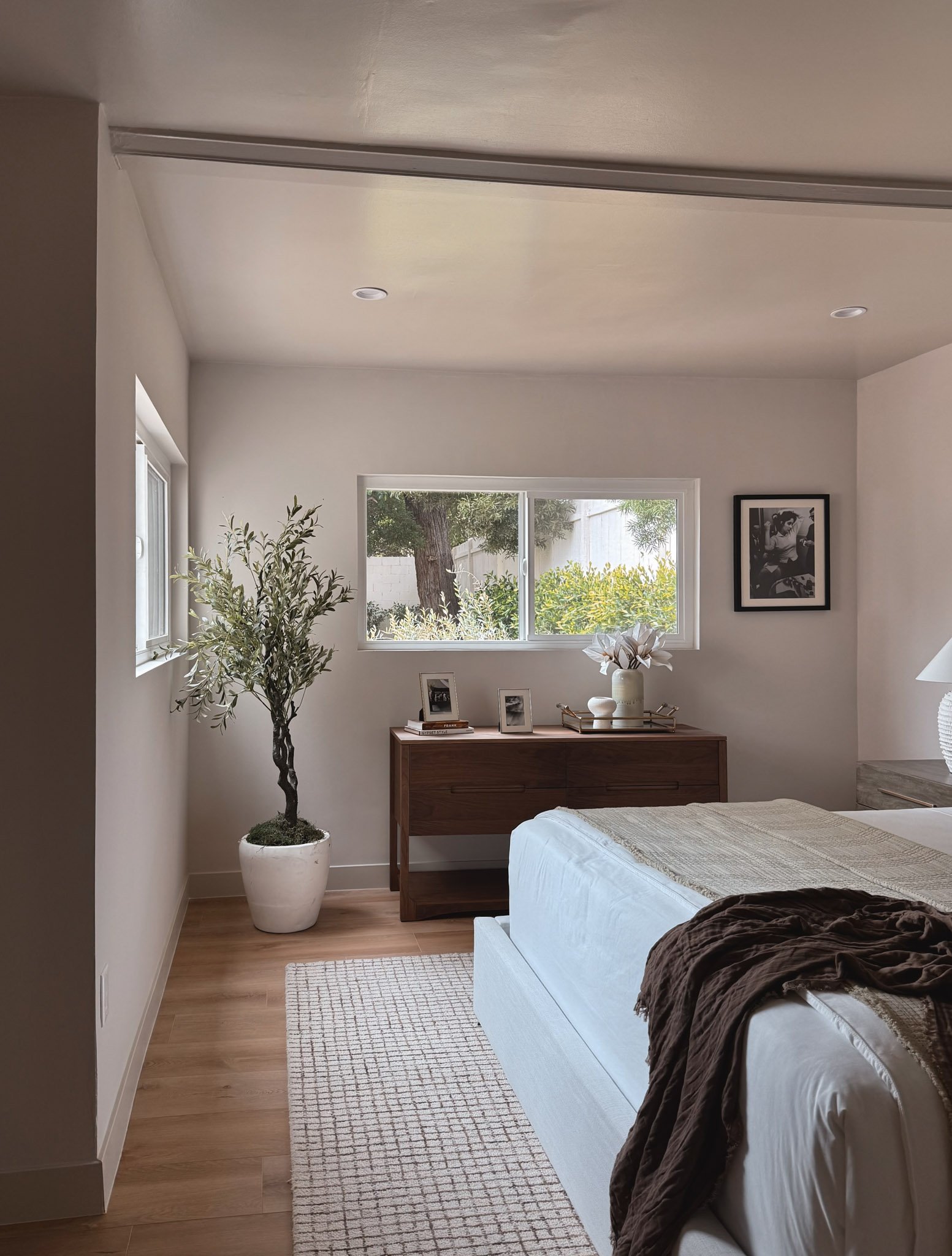 A bedroom with a bed, a wooden dresser with photo frames and a vase, a large potted plant, and windows showing outdoor greenery.