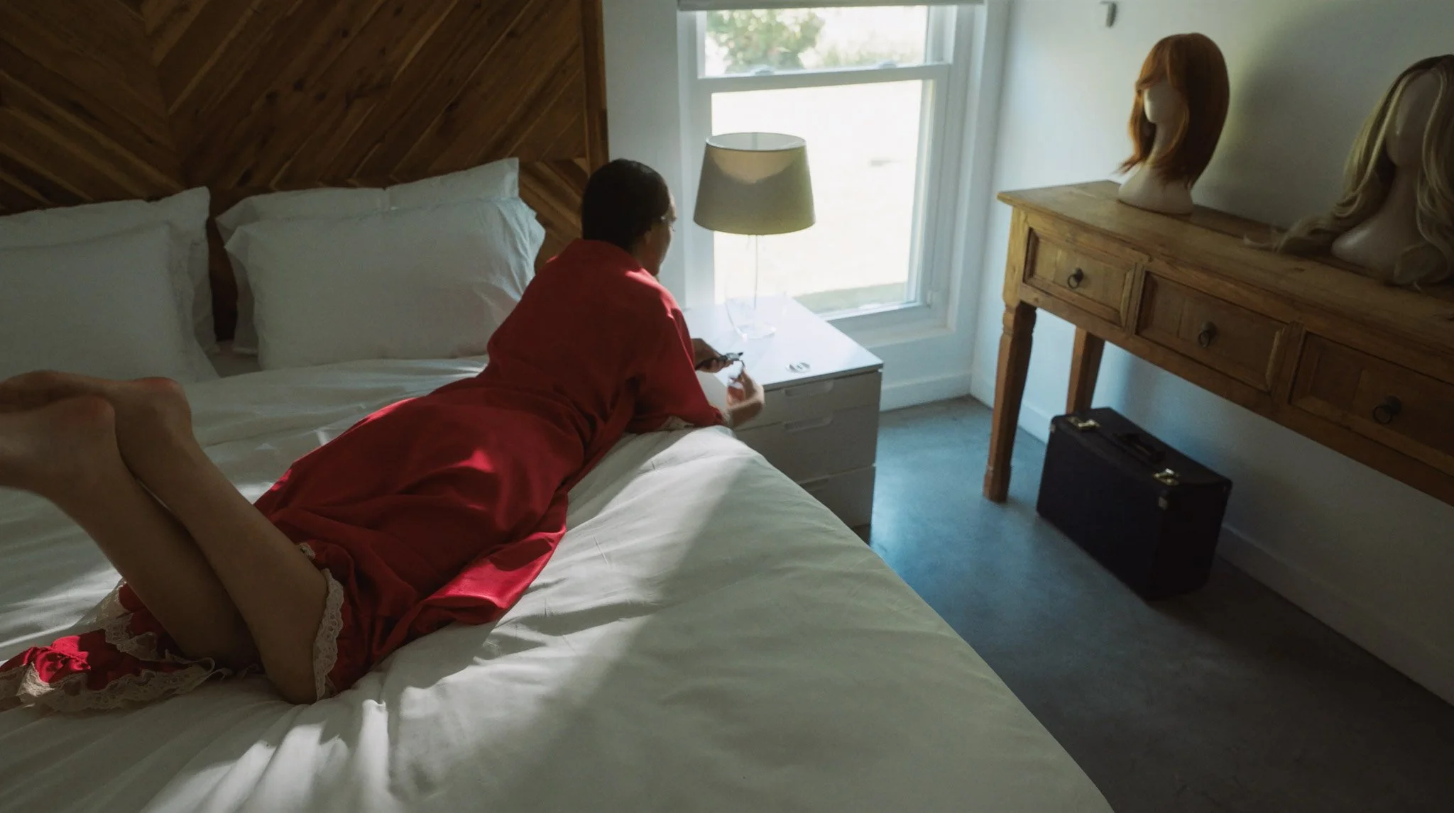 A woman in red pajamas with lace trim lying on her stomach on a bed, looking at her phone in a bright bedroom with a wooden headboard, white pillows, and a nightstand with a lamp. There are mannequin heads with wigs on a wooden dresser near a large window.