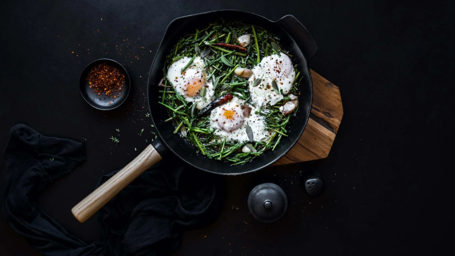 A skillet with cooked greens topped with three poached eggs, garnished with herbs and seasonings, placed on a wooden board on a dark surface.
