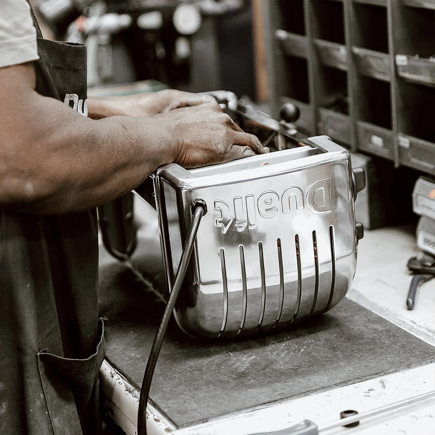 A person with dark skin wearing a black apron moves a stainless steel toaster on a worktable in a workshop or kitchen setting.