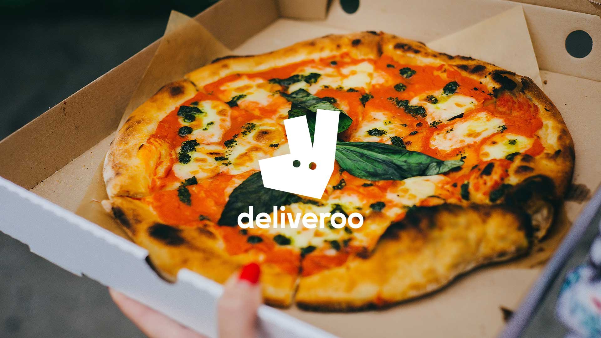 A pizza with cheese, spinach, and tomatoes in a delivery box, with the Deliveroo logo overlay.