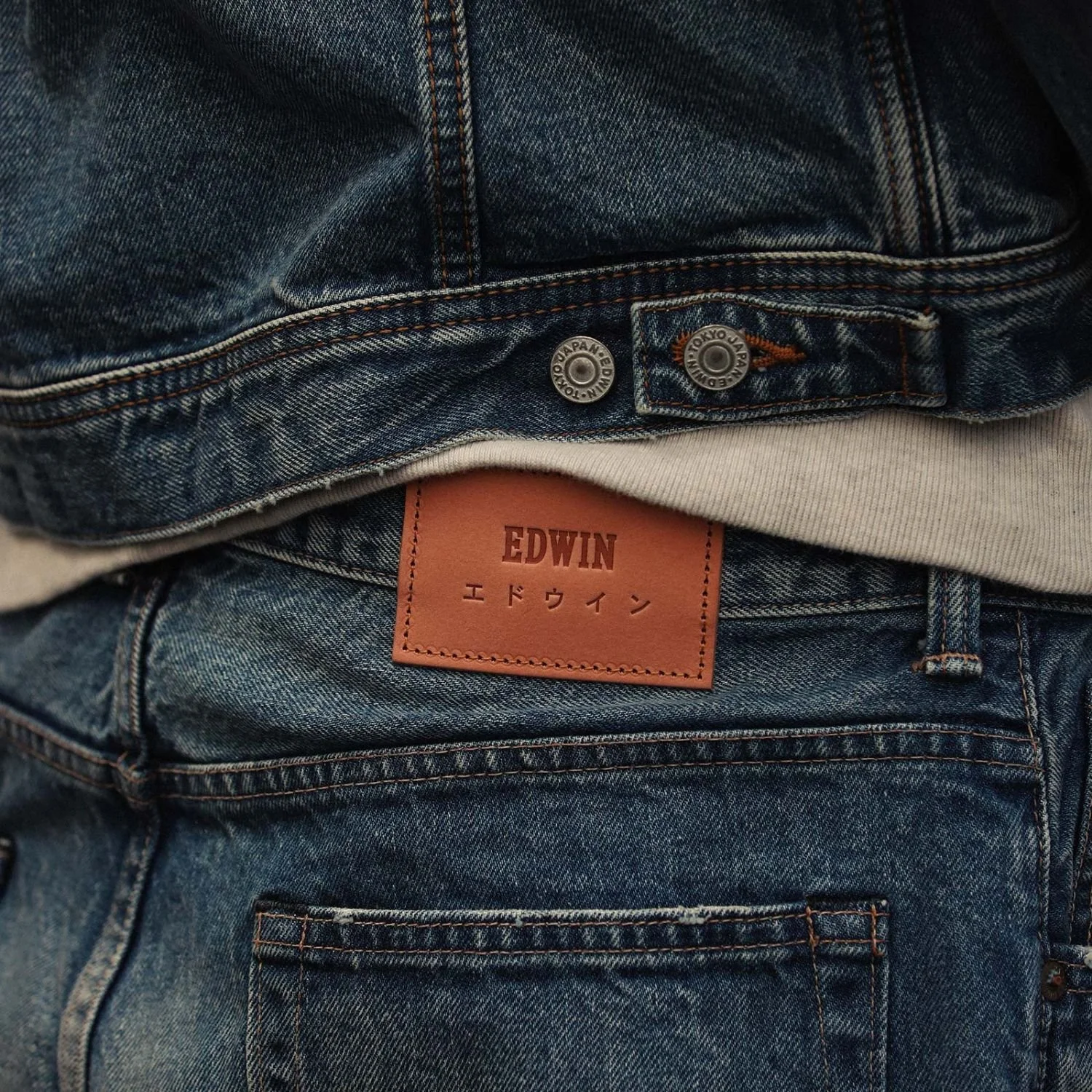 Close-up of a stack of blue denim jeans and a jacket showcasing a tan label with the brand name 'EDWIN' and Japanese characters, partially visible, along with the waistband of the jeans.