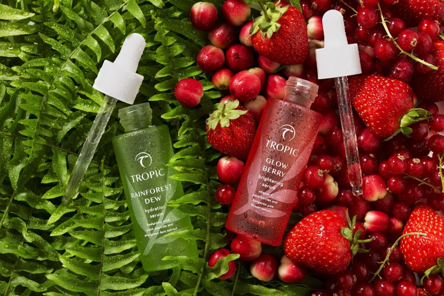 Two bottles of Tropic skincare serums, one labeled Rainforest Dew and the other Glow Berry, placed among ripe strawberries and red berries on a bed of green fern leaves with water droplets.