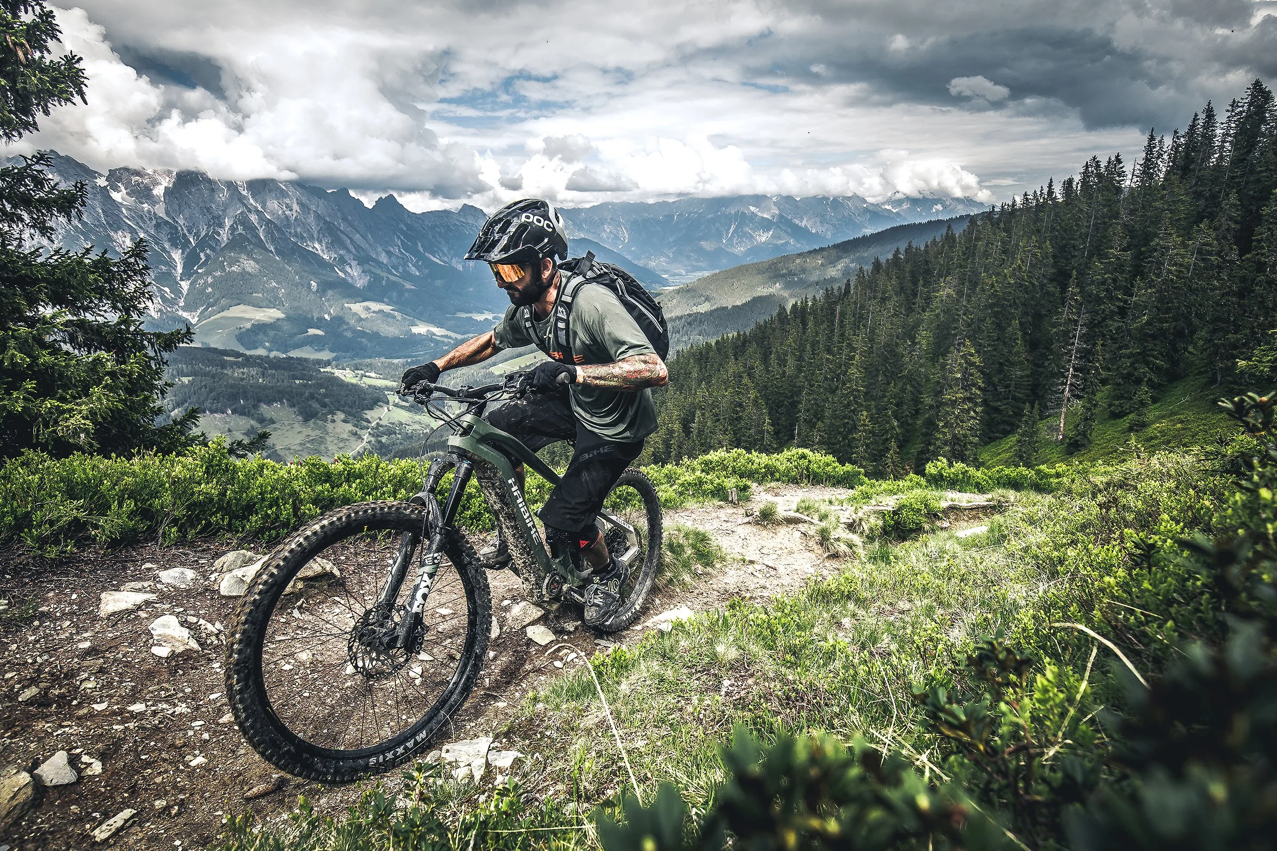 A mountain biker wearing a helmet, sunglasses, and riding gear navigates a dirt trail through lush green terrain with mountainous landscape and cloudy sky in the background.