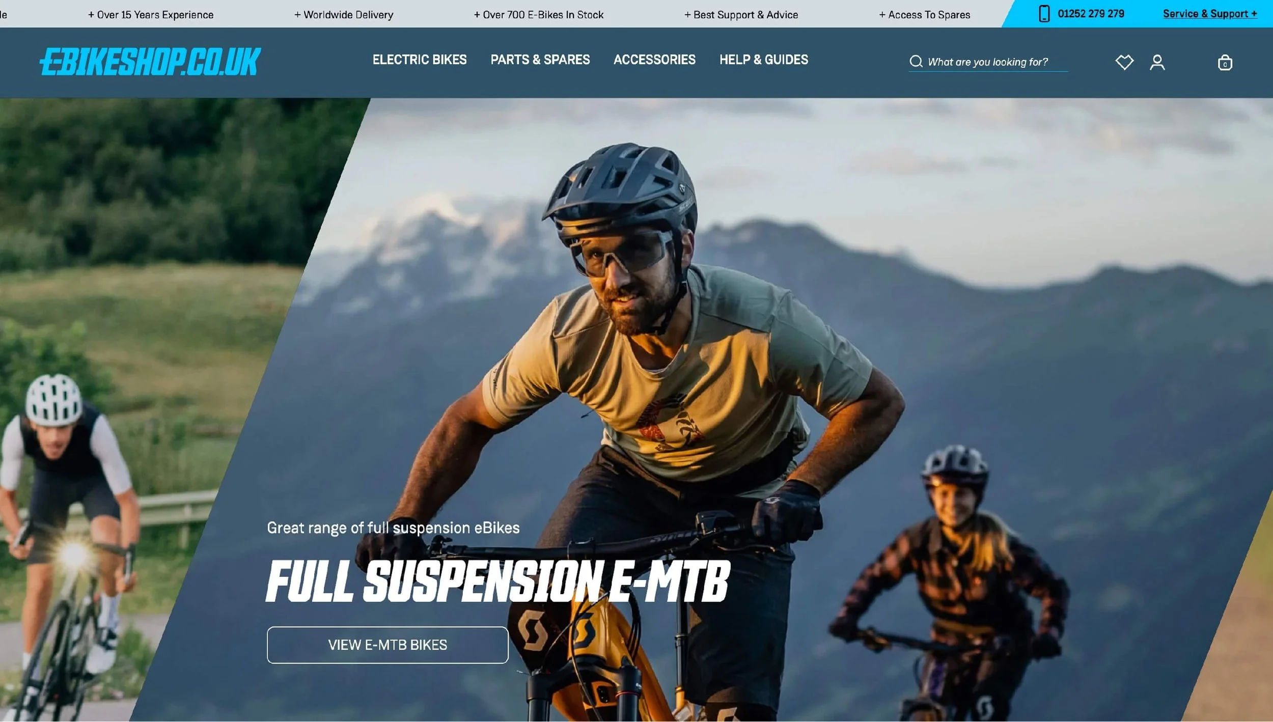 Homepage of ebikeshop.co.uk showcasing an outdoor scene with mountain bikers, featuring a man in a helmet and sunglasses riding a full suspension e-MTB, with a mountain range in the background.