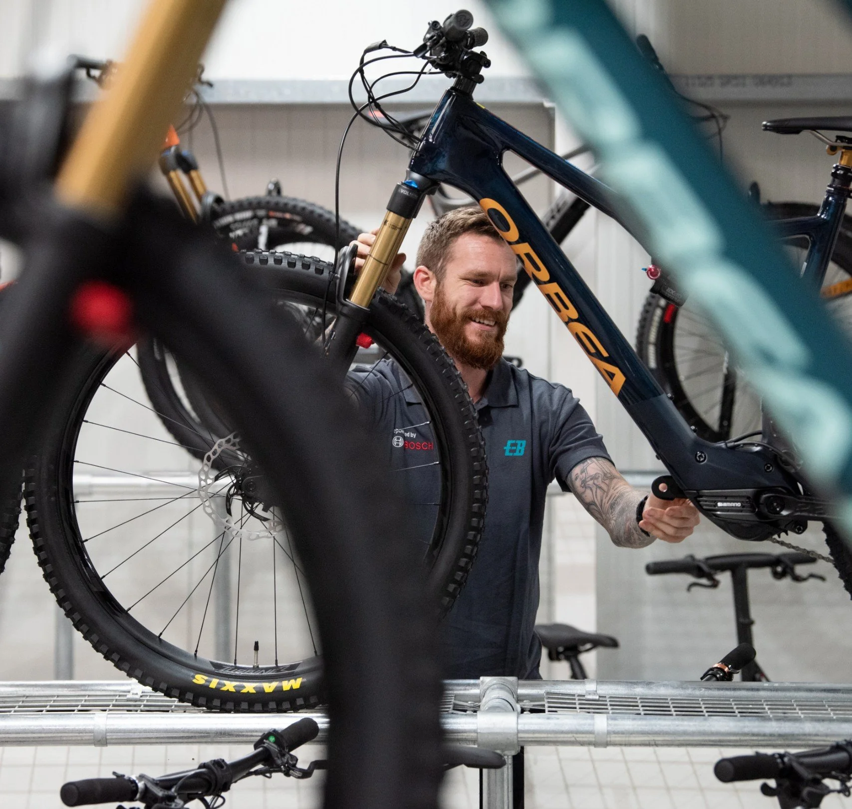 A man with a beard and tattooed arm working on a blue and gold mountain bike in a workshop, surrounded by other bicycles and bike parts.