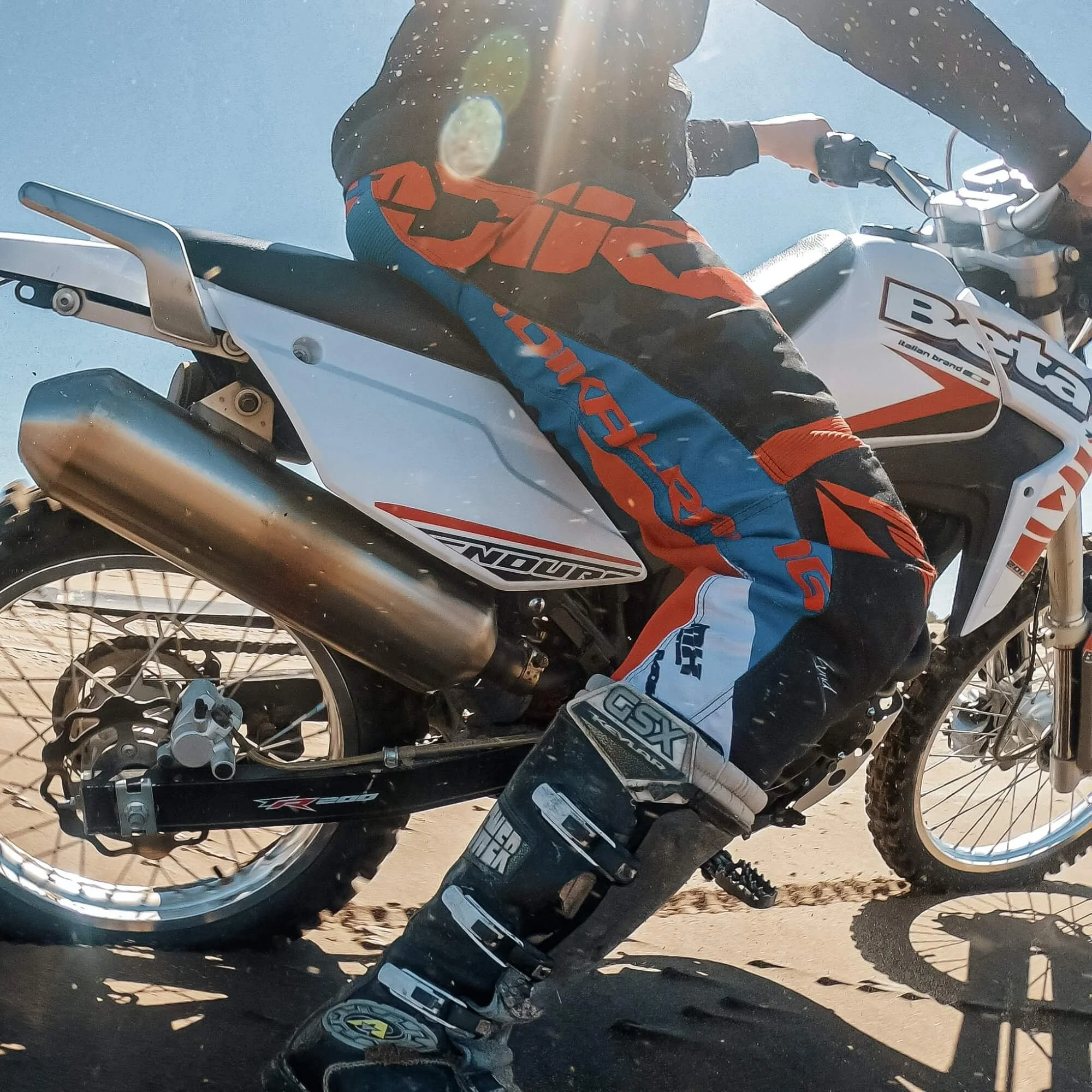 Close-up of a motocross rider sitting on a white and black dirt bike with orange and blue riding gear, on a dirt track under sunlight.