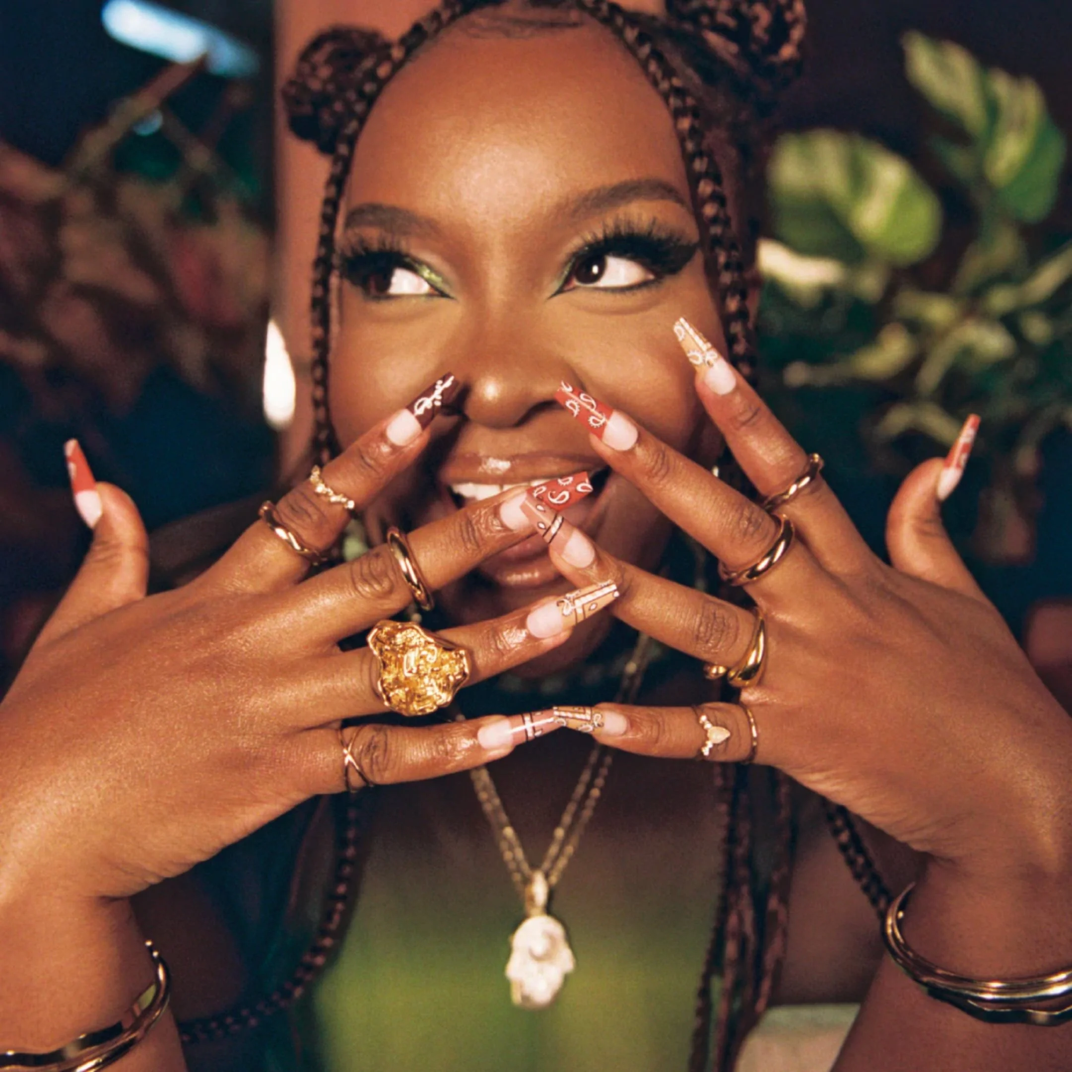 Close-up of a woman with braids, wearing jewelry and colorful manicured nails, smiling and making a playful gesture with her hands around her face.