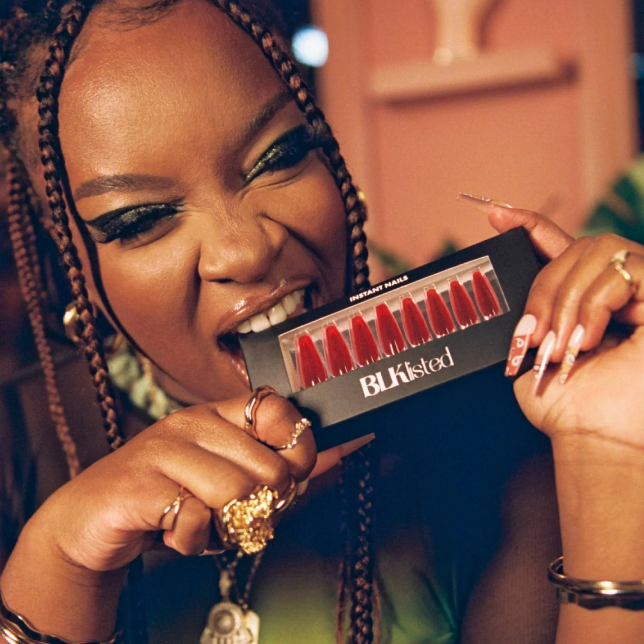 A woman with braided hair and gold jewelry biting into a box of red lip glosses labeled 'BLK Lusted'.