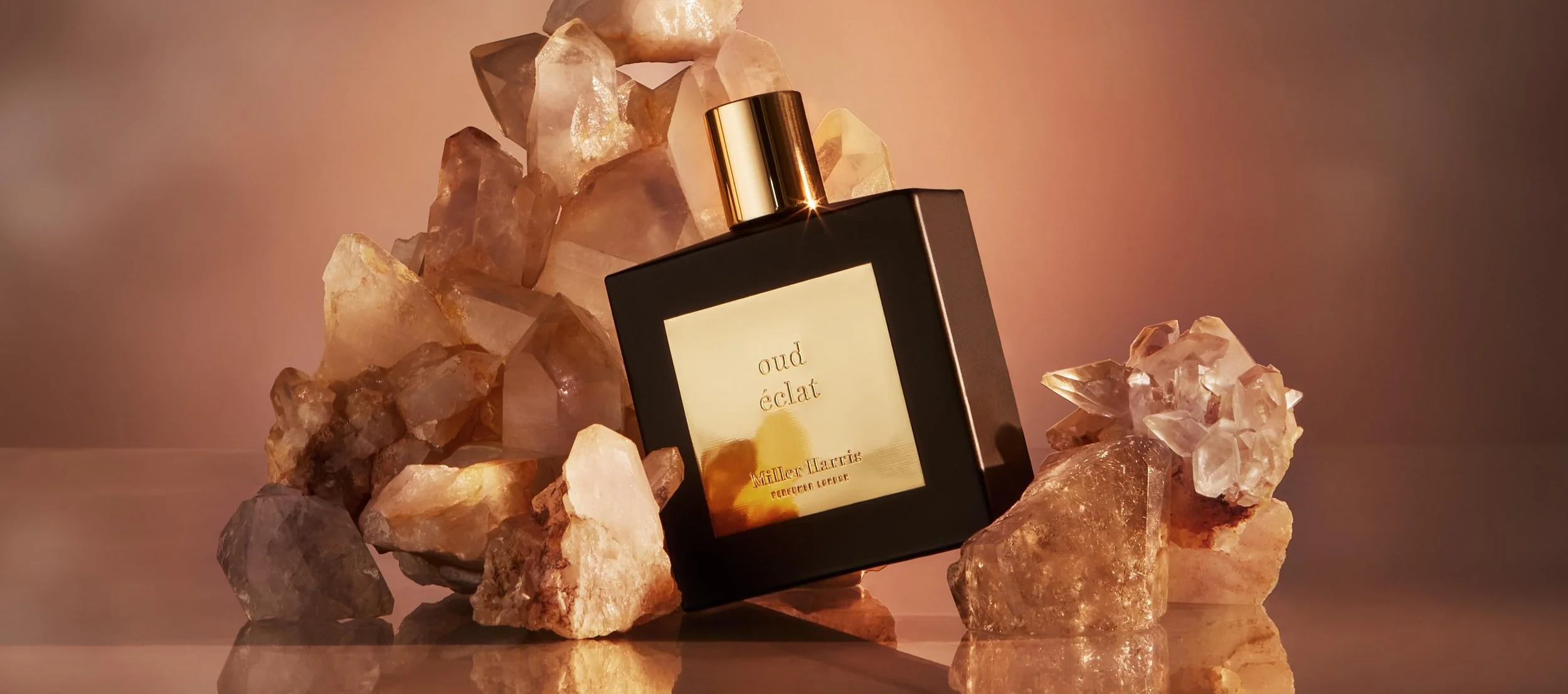 A black perfume bottle labeled 'oud éclat' by Miller Harris is surrounded by various translucent and opaque crystal rocks, with a warm, soft lighting atmosphere.