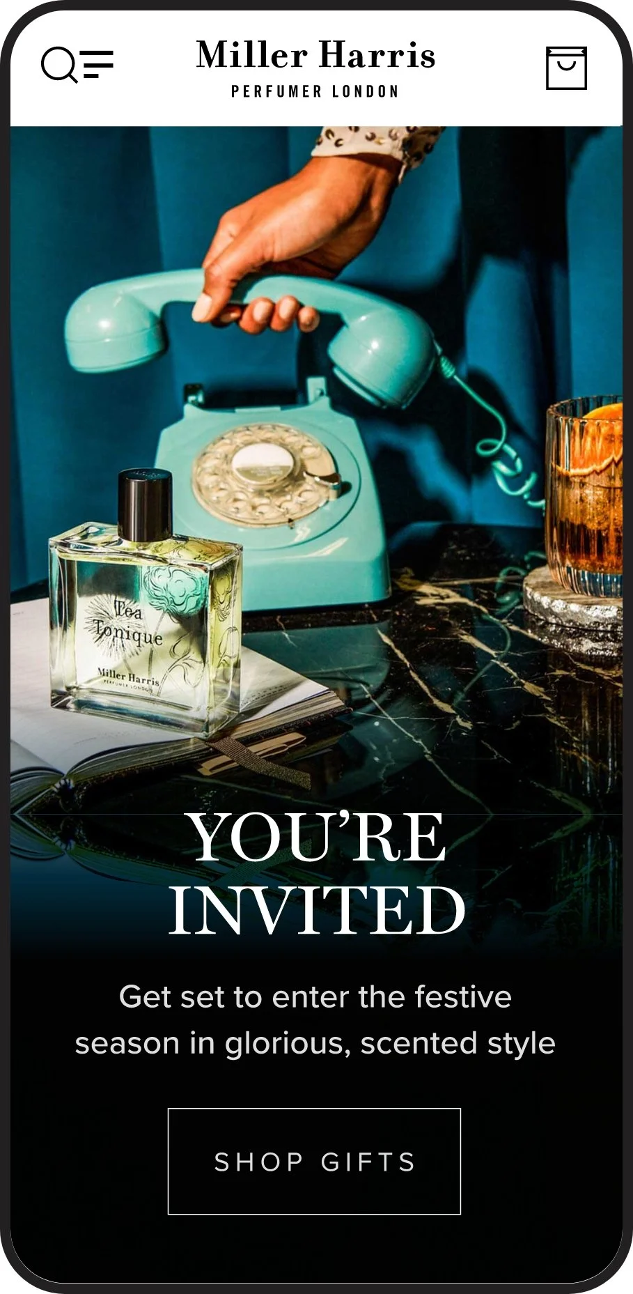 A promotional graphic for Miller Harris featuring a vintage teal rotary phone, a bottle of perfume labeled 'Tea Tonic,' a notebook with a pen, a glass of amber-colored drink, and a candle on a black marble surface. The text invites viewers to enter a festive season and shop gifts.