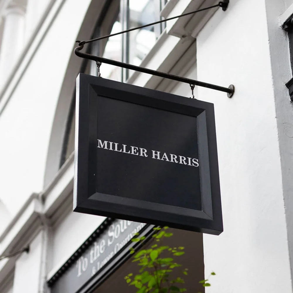 A black square sign with white text reading "MILLER HARRIS" hanging from a black metal rod outside a building, with a window and part of another sign visible in the background.
