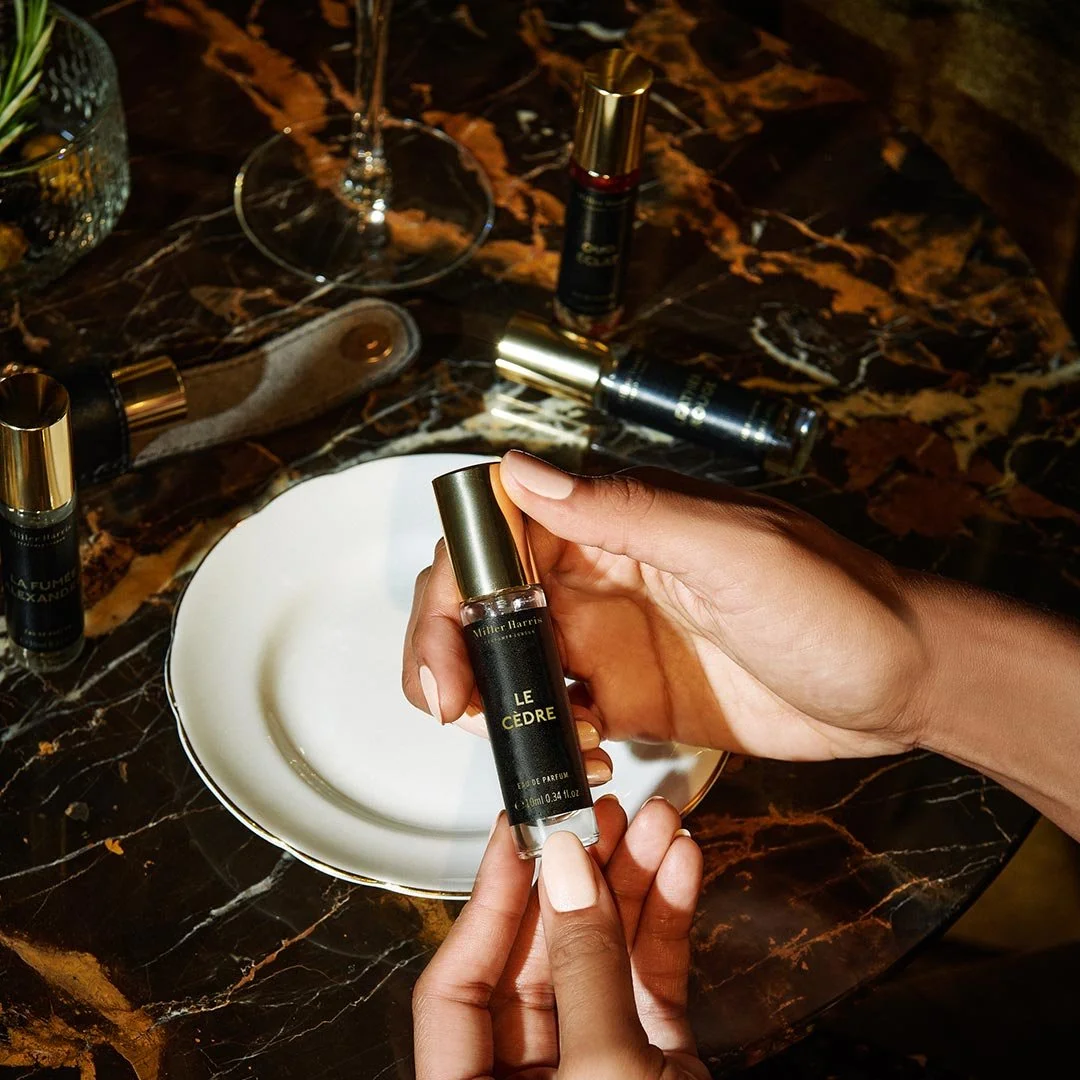 A woman holding a perfume bottle labeled "Le Cèdre" over a white decorative plate on a dark marble table, with several other perfume bottles and a wine glass in the background.