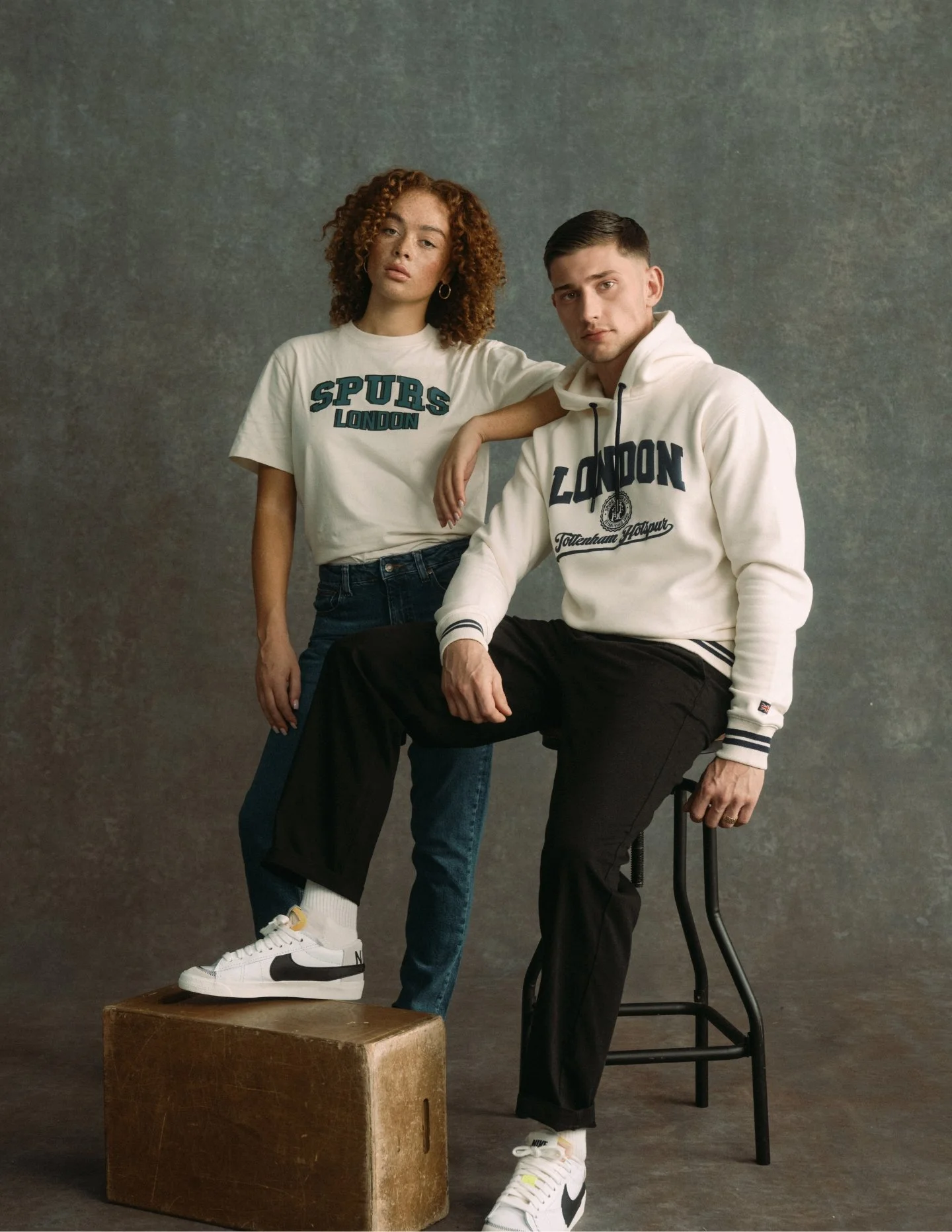 Two young adults, a woman and a man, pose against a plain background. The woman stands with her left hand resting on the man's shoulder, wearing a white t-shirt and blue jeans. The man sits on a black chair with his right foot on a wooden box, wearing a white hoodie and black pants.