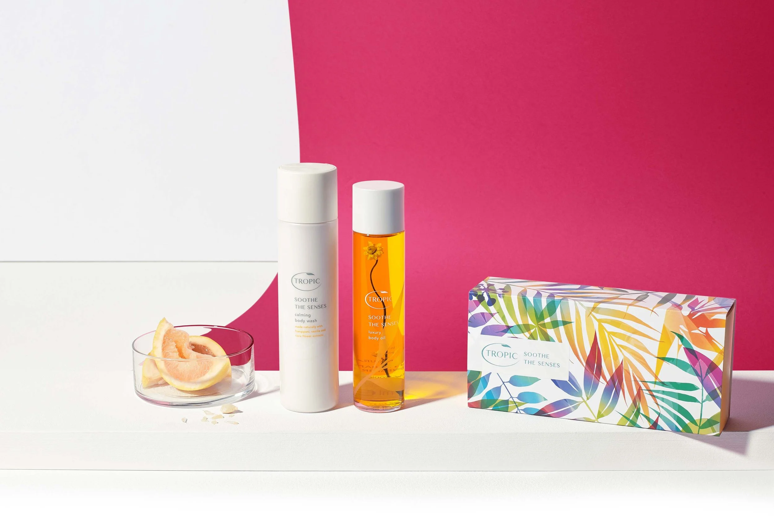 Beauty products including a white bottle, an orange body oil, a floral-themed box, with a plate of grapefruit slices on a white surface against a pink and white background.
