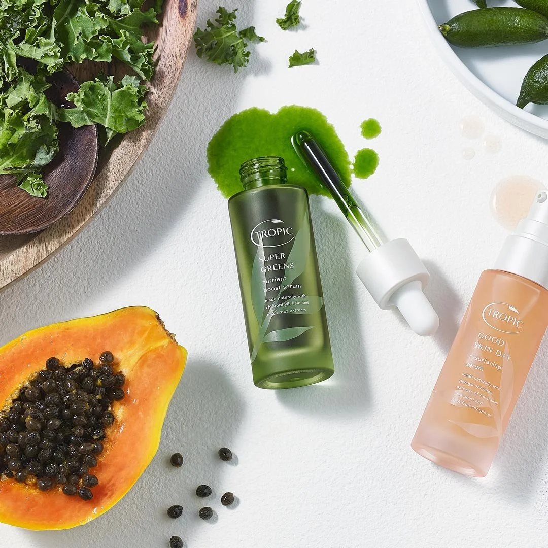 Flat lay of skincare products among fresh fruits and greens, including a papaya half with black seeds, a bowl of mixed greens, and greenery on a white surface.