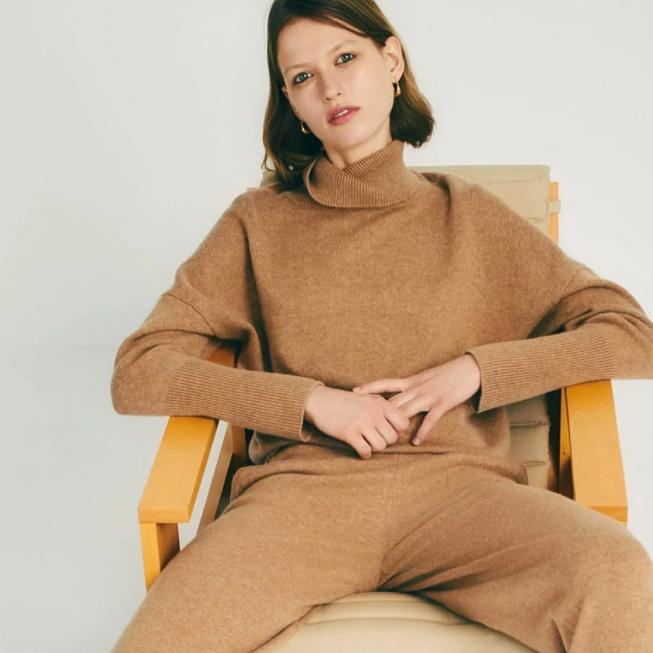A woman with short brown hair and earrings sitting in a wooden chair wearing a brown turtleneck sweater and matching pants.