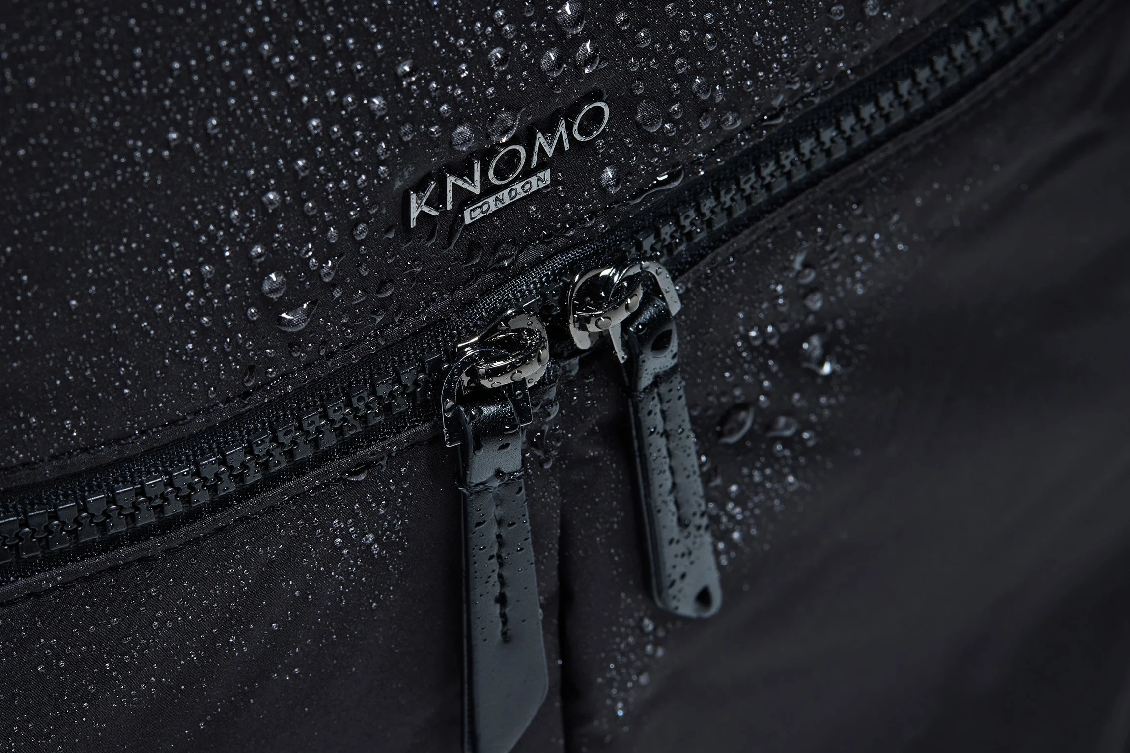 Close-up of a black Knomo London bag with water droplets on its surface and zippers.