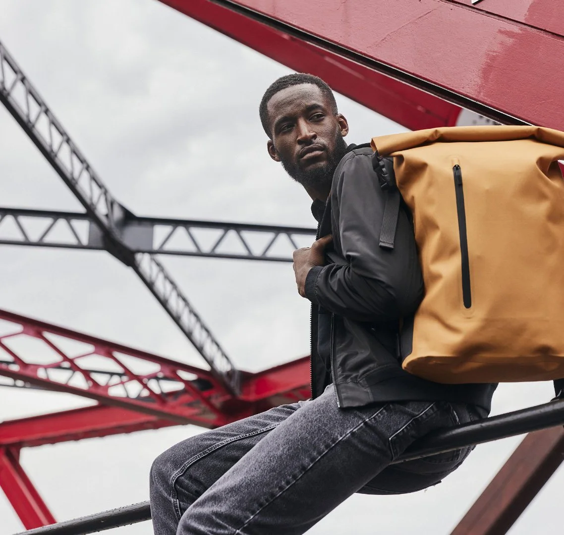 A young man with dark skin and short hair carries a tan backpack while sitting on a metal beam, with a red and black industrial structure in the background.