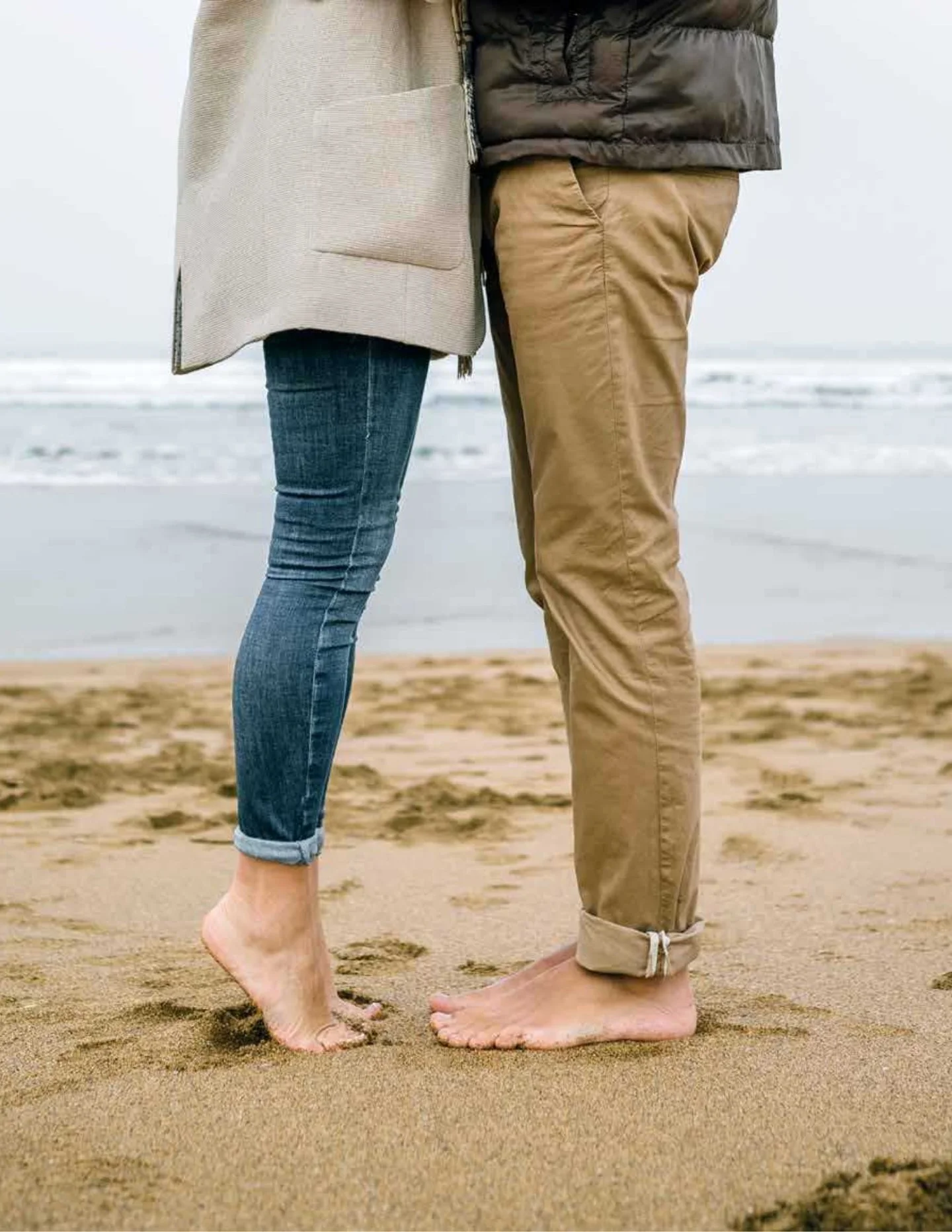 Close-up of a couple standing barefoot on the beach, showing their legs and feet, with the ocean and cloudy sky in the background.
