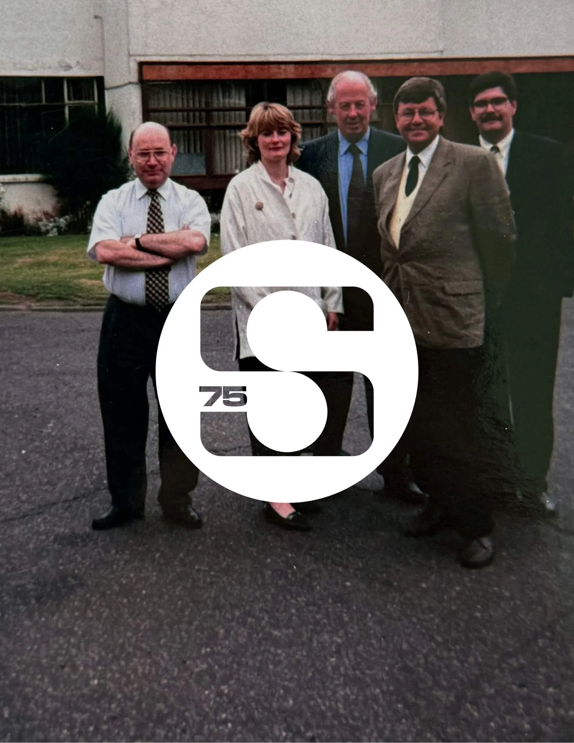 A vintage photograph of five people standing outdoors in front of a building with greenery, with a large logo with the number '75' and the letter 'S' overlapping the bottom center of the photo.
