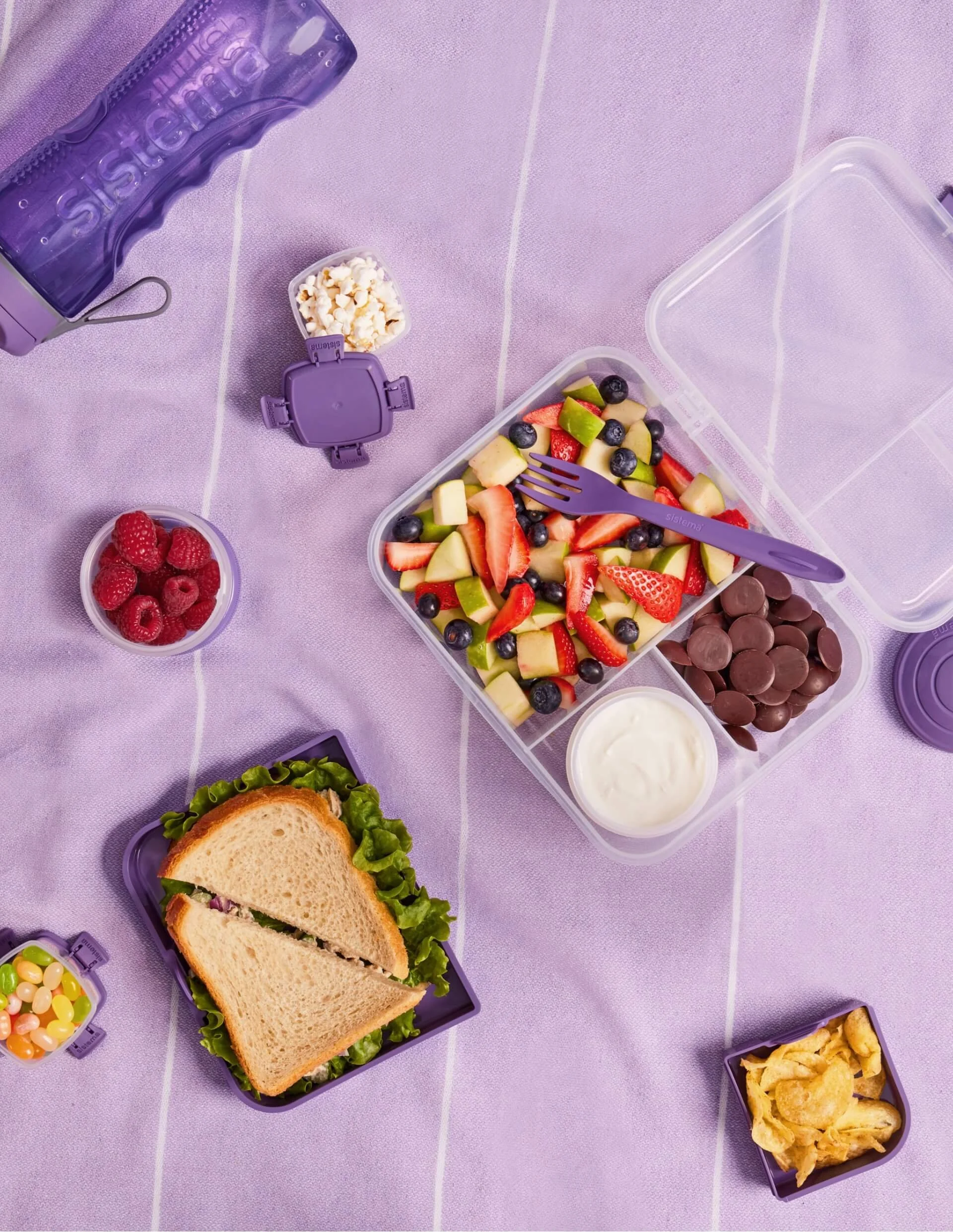 A lunchbox with fresh fruit salad, chocolate candies, a sandwich, chips, and small containers of popcorn, jelly beans, and yogurt on a purple tablecloth.