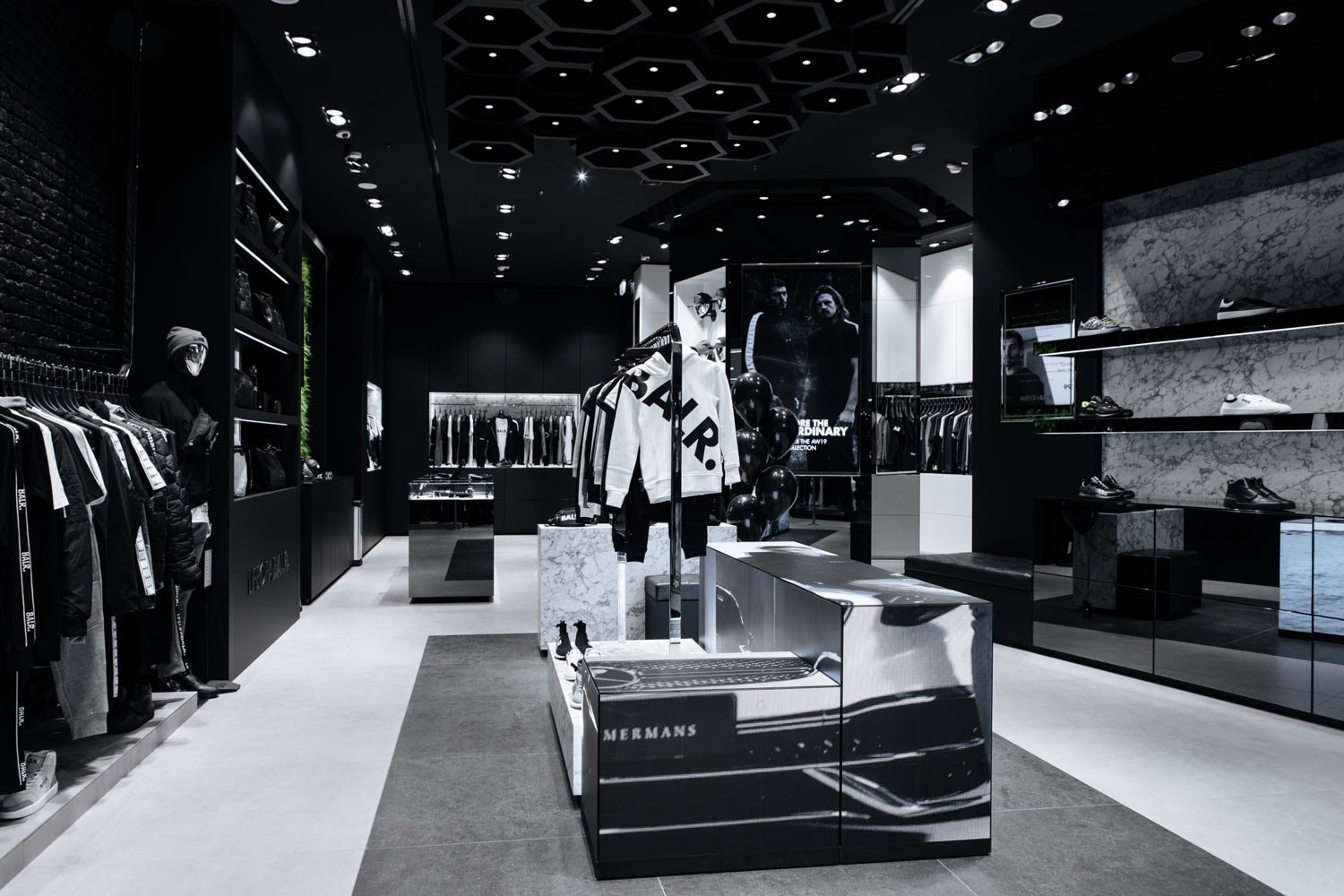 Interior of a modern clothing store with black walls and ceiling, featuring racks of clothes, shelves with shoes, and decorative elements in black, white, and gray tones.