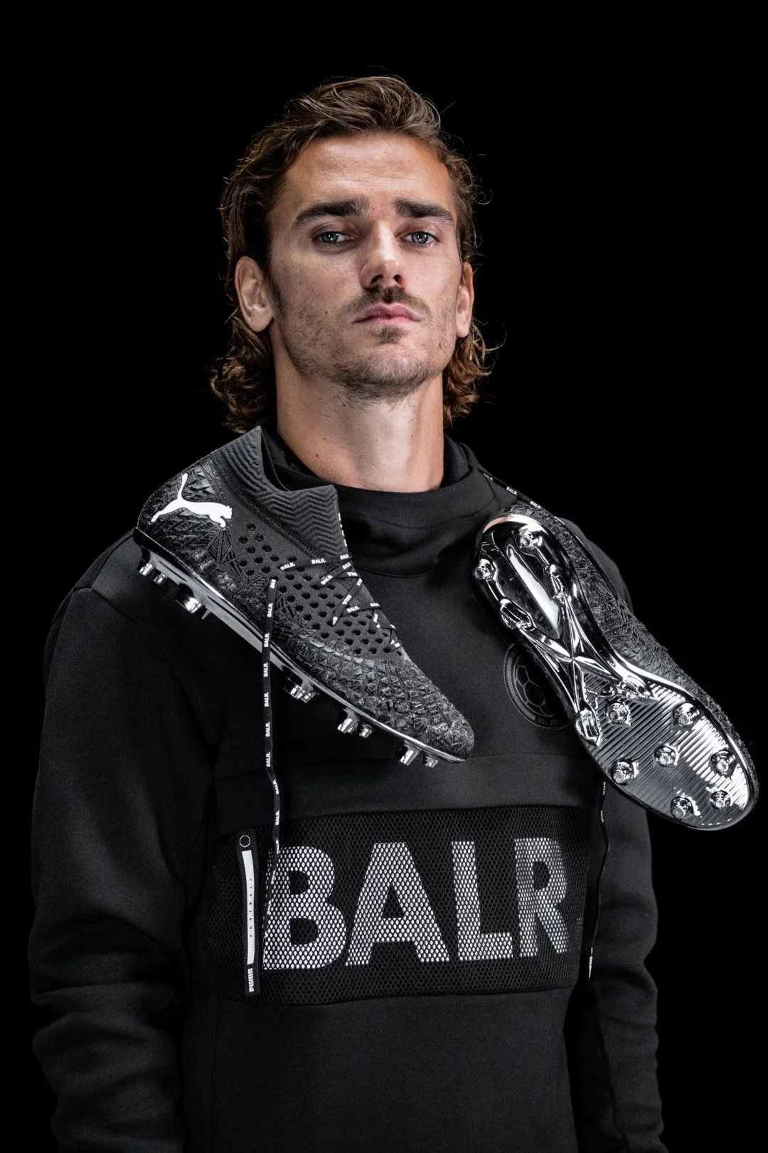 A man with long, wavy brown hair and a serious expression is wearing a black sports outfit with the word 'BALR' on it. He has two black and silver football cleats draped over his shoulders.
